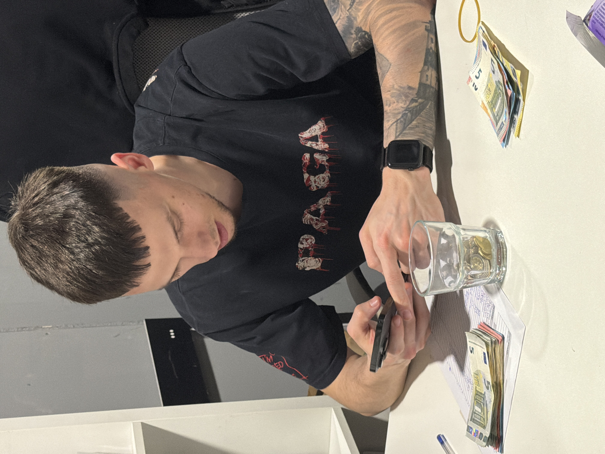 A medium shot depicts a light-skinned male with dark hair and visible tattoos on his left forearm. He is wearing a black t-shirt with red and white lettering. He is seated at a white desk and holding a black smartphone in his left hand, with his right hand resting near a glass filled with coins. A stack of Euro banknotes lies on a paper below the glass. Another stack of banknotes is visible in the upper right corner of the frame, secured with a yellow rubber band. The setting appears to be indoors. The location is unknown.