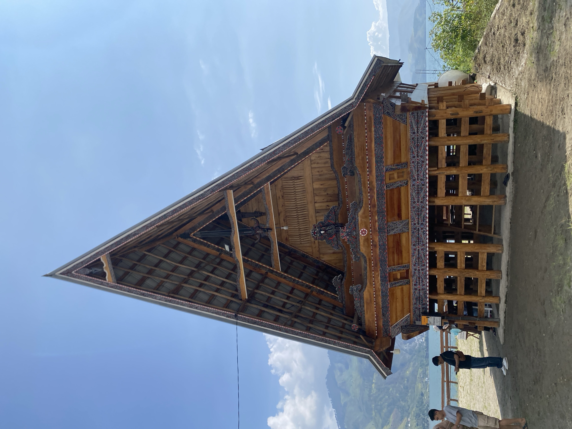 The image shows a traditional wooden stilt house with a prominent triangular roof in Parlondut, Indonesia. The house is ornately decorated with intricate patterns in red, black, and white along the eaves and gable. The roof is made of corrugated metal. The structure is elevated on sturdy wooden posts and appears to be situated on a slight incline overlooking a body of water, likely a lake, with rolling hills and mountains in the background. The sky is a clear blue with a few scattered white clouds. In the foreground, two men are standing and looking at their phones. Another person, partially obscured, is also visible near the house. The ground around the house is a mixture of dirt and dry grass. A wooden fence runs along the edge of the property.