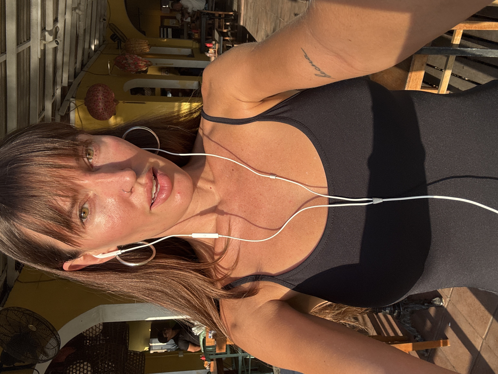 A woman with long brown hair and bangs, wearing a black tank top and hoop earrings, takes a selfie in what appears to be an outdoor restaurant in North Kuta, Indonesia. She has white earbuds in her ears and a small nose ring. Sunlight casts strong shadows on her skin, highlighting her freckles and tanned complexion. A script tattoo is visible on her left arm. In the background, a ceiling fan is mounted on a yellow wall, and decorative lanterns hang from the ceiling. Several tables and chairs are set up, and other patrons can be seen in the distance. The overall ambiance suggests a warm, sunny day in a tropical setting.