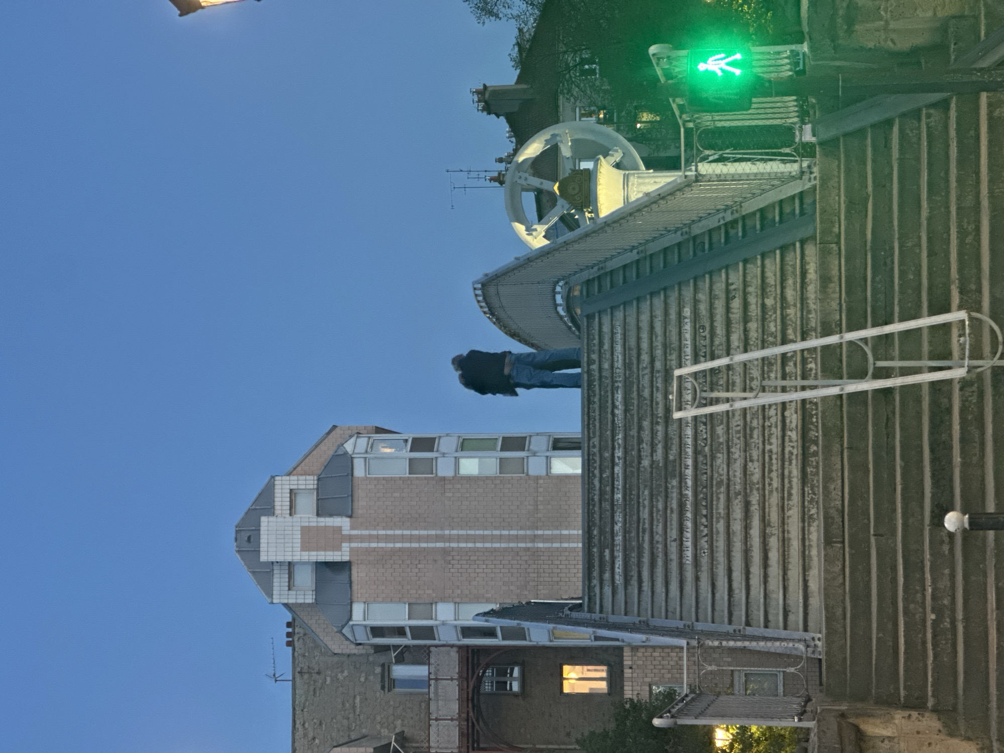 The image depicts a cityscape in Paris, France, at dusk. The sky is a clear blue. A building with a unique roof design, featuring light-colored bricks and multiple windows, is visible. A person in dark clothing and blue jeans is standing on a corrugated metal roof near an object with a rounded, mechanical appearance. In the background, a pedestrian crossing sign shows a green walking figure. A series of concrete walls or barriers is on the right side.