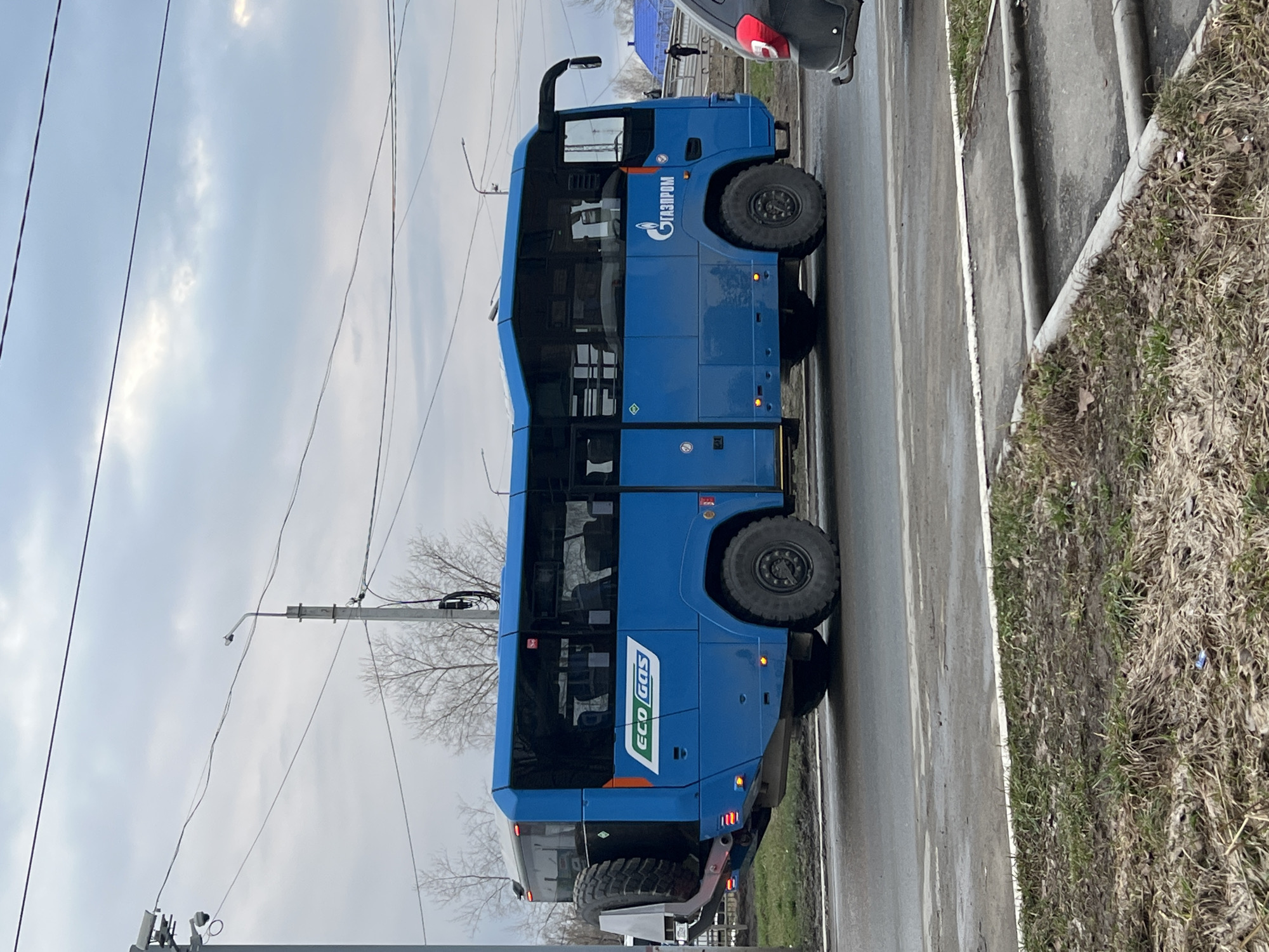 A blue GAZprom EcoGas bus is parked on the right side of a wet road in Chaikovsky, Russia. The bus has large, off-road tires. The sky is overcast. To the right of the road is a concrete curb and an area of dry grass. Part of a grey car is visible behind the bus. A power pole is visible above and behind the bus.