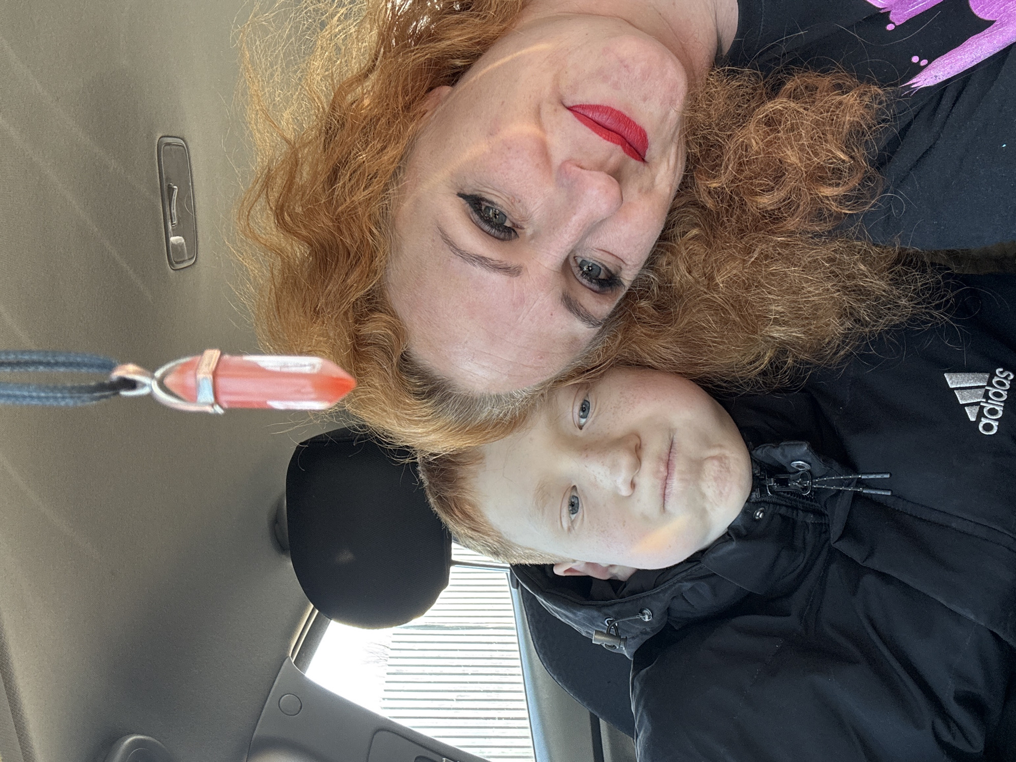 A selfie taken inside a car shows an adult female and a child. The adult female has red curly hair, red lipstick, and eyeliner, wearing a dark shirt with purple designs. She is positioned above the child. The child, presumably a boy, also has red hair and freckles and wears a black Adidas jacket. A red crystal pendant hangs in the foreground. The car interior includes a gray roof, a black headrest, and part of the dashboard with car features. There is no information available to pinpoint the exact location in Chaikovsky, Russia.