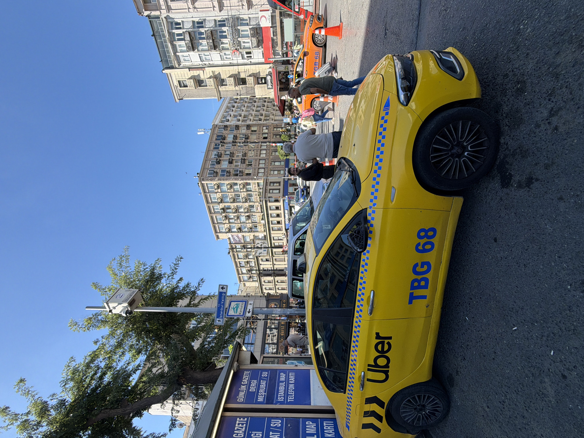 A sunny day in Istanbul, Turkey, where a yellow Uber taxi with the license plate "TBG 68" is parked on a city street. The taxi has a blue and white checkered stripe along its side and a distinct Uber logo. Several people are visible in the background, some standing and conversing near a newsstand, while others are walking across the street. An orange taxi is also present further down the road, along with traffic cones. The urban environment is characterized by multi-story buildings and trees. A traffic camera mounted on a pole is also visible.