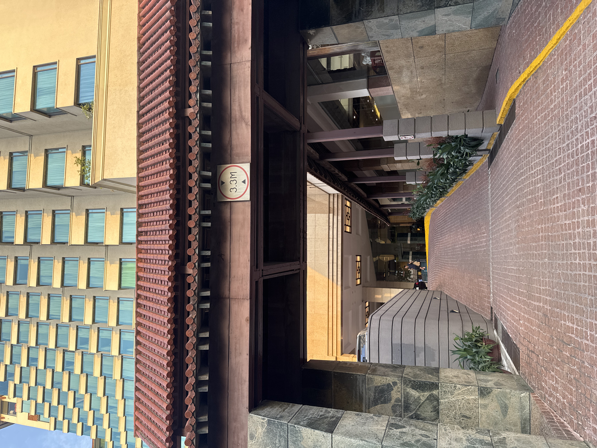The image shows the entrance to a building with a sloped driveway paved with red cobblestones. A sign indicating a height limit of 3.3 meters is visible on a wooden beam above the entrance. The building has a modern facade with many windows and is partially obscured by a traditional-style tiled roof overhang. Green plants are visible in planters along the side of the driveway. The scene appears to be in an urban environment, likely in Hà Nội, Vietnam, given the architectural style and context. A few people are visible in the background, near the entrance of the building, and a white car is partially visible on the driveway. The lighting suggests it is daytime, with sunlight casting shadows.