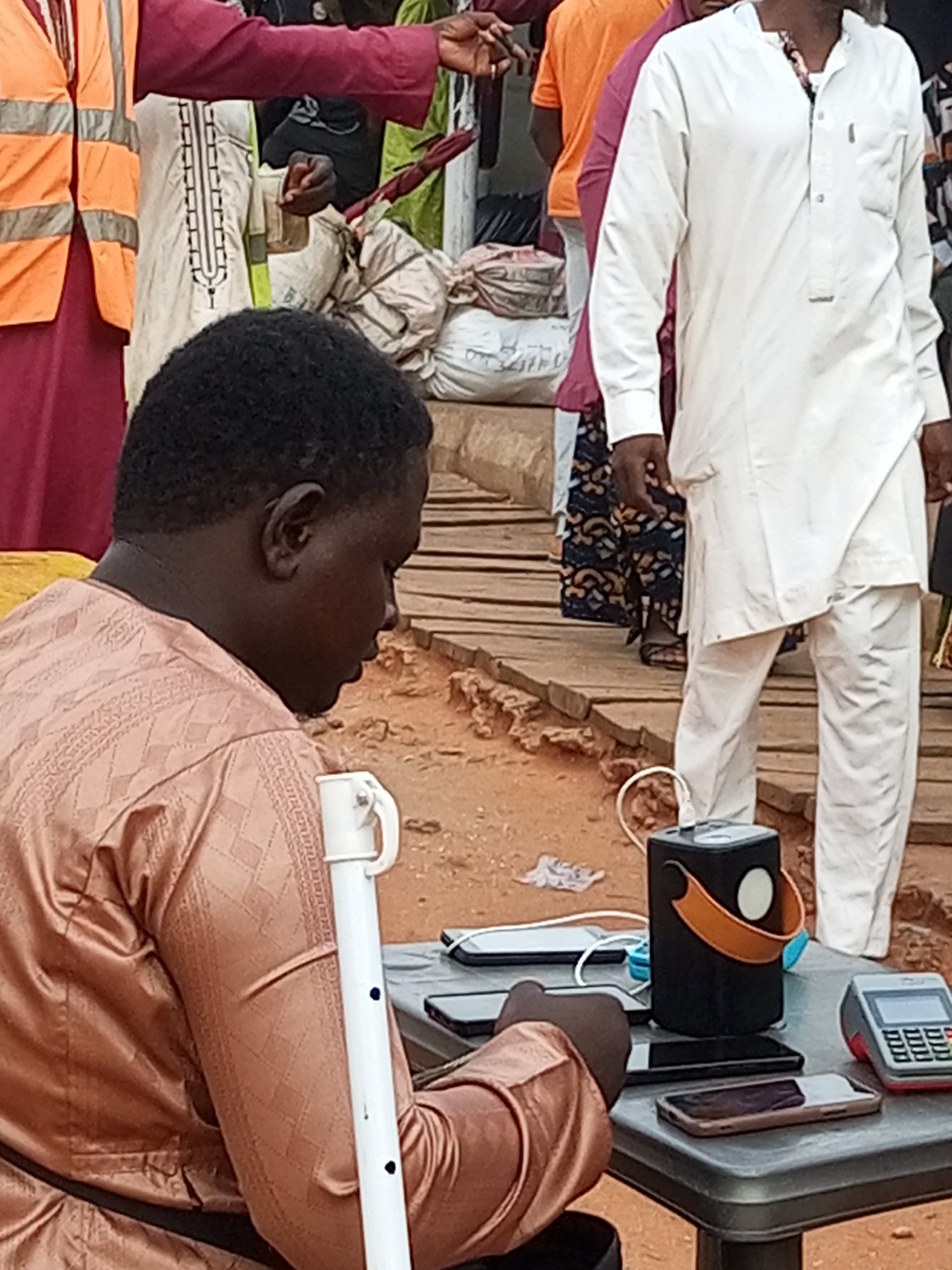 The image depicts a street scene in Gombe City, Nigeria. In the foreground, a person wearing a patterned brown garment is seated and looking down at a table. On the table are several mobile phones, a portable speaker, and a point-of-sale device. Behind the seated individual, other people are visible, some wearing traditional attire. An individual in a white outfit is standing and facing away from the camera. Another person is wearing an orange t-shirt. The ground appears to be dirt, and there are wooden planks in the background. The overall setting suggests an outdoor market or a bustling urban area.