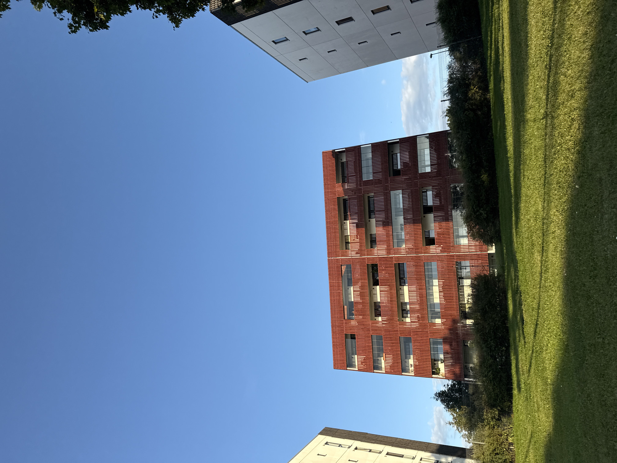 The image shows a clear blue sky above two apartment buildings and a grassy lawn in the foreground. One building is predominantly red with a patterned facade and balconies, while the other is white with a more minimalist design. A third building, partially visible on the left, is light-colored with vertical windows. Green bushes and a fence are situated between the buildings and the lawn. The scene appears to be in a residential area, likely in the city of Tallinn, Estonia, given the architectural style and typical urban greenery. No people or specific actions are visible.
