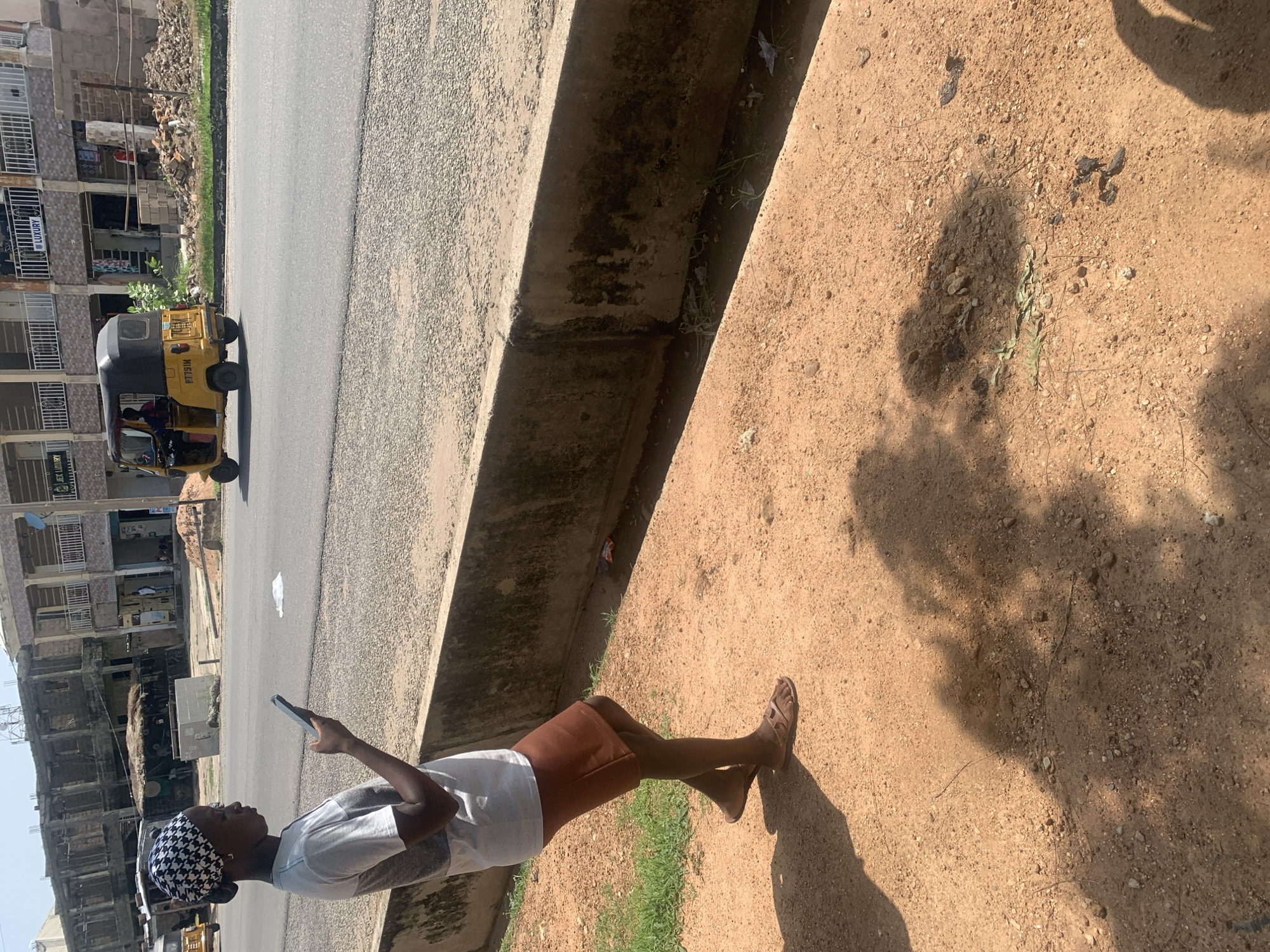 In Jalingo, Nigeria, a young person stands on a dusty, sun-drenched ground, holding a smartphone. They are dressed in a white shirt and a brown skirt, with a black and white patterned headscarf. Their attention is focused on the phone, suggesting they are engaged with its content. The scene is set next to a road, where a yellow auto-rickshaw is visible, suggesting local transportation.
Behind the auto-rickshaw, buildings line the street, some appearing to be under construction. The architecture hints at a developing urban environment. The sunlight casts strong shadows on the ground, indicating it is likely midday or early afternoon. The overall impression is a snapshot of everyday life in a Nigerian town, with the individual absorbed in their digital world amidst the hustle and bustle of their surroundings.
The image captures a moment of quiet personal engagement against a backdrop of urban activity and construction. The bright sunlight and dusty terrain are characteristic of the region. No specific emotional cues are discernible from the person's posture or expression. The text "LUXURY" is visible on a sign on one of the buildings.