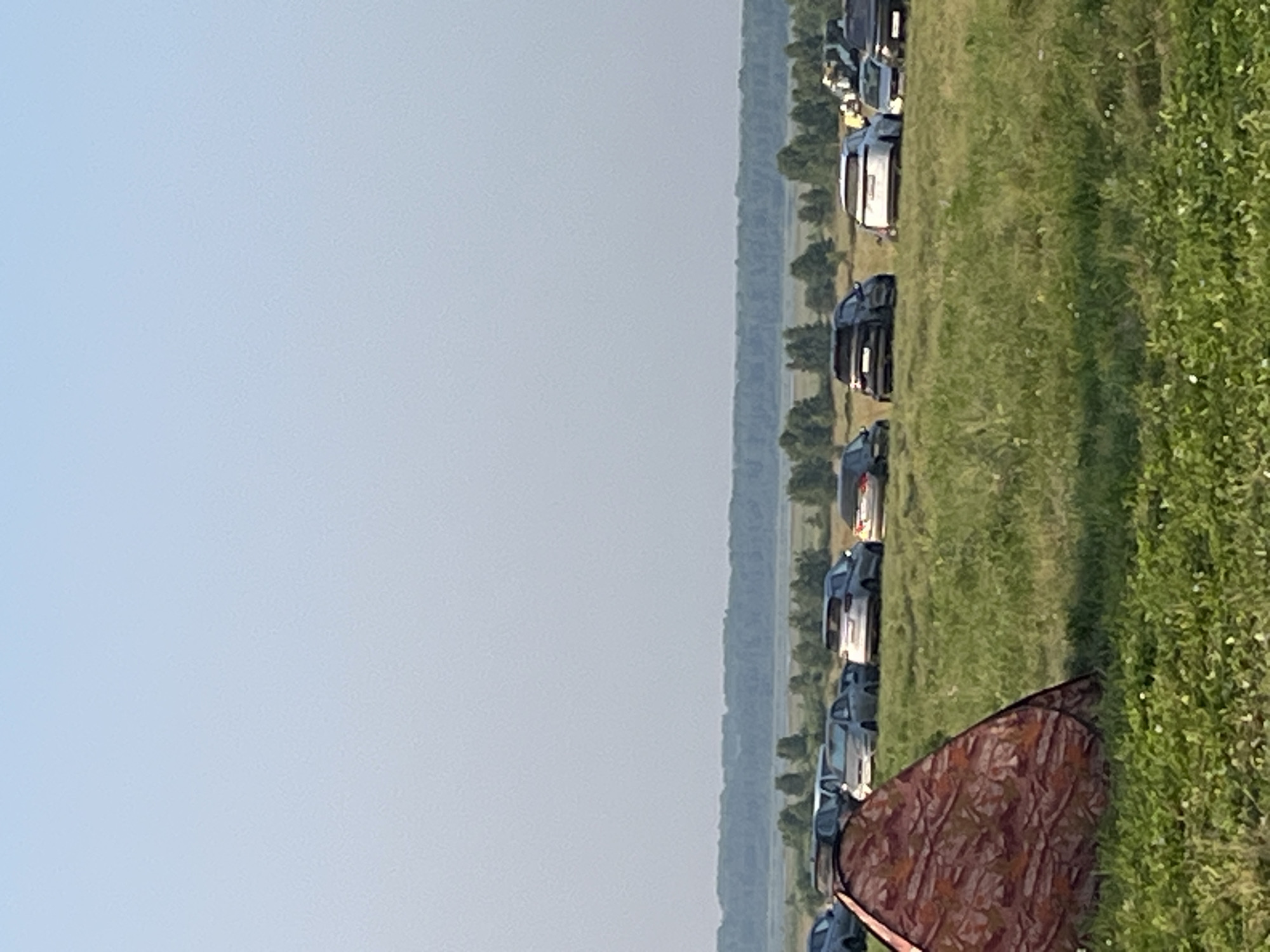 In Surakovo, Russia, a camouflage tent is pitched in a grassy field with a clear blue sky overhead. A line of parked cars, including a white SUV, a black SUV, and several other sedans and SUVs, are visible in the middle ground. Beyond the cars, a dense line of trees forms the horizon, suggesting the edge of a forest or park. The overall scene depicts an outdoor gathering or event taking place in a rural or semi-rural setting.