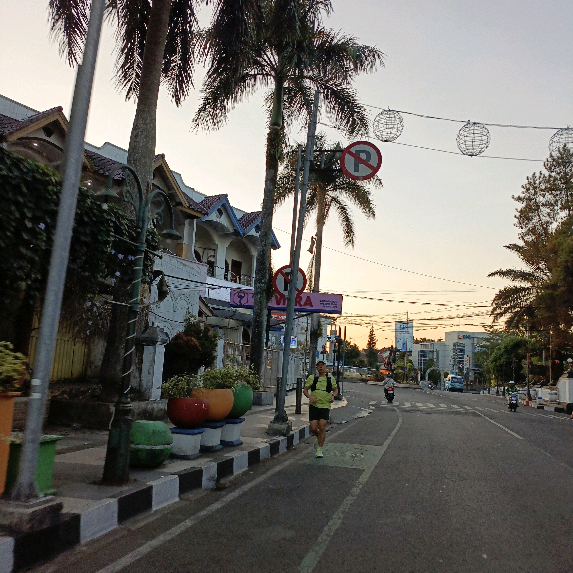 The image depicts a street scene in Salatiga, Indonesia, during what appears to be sunrise or sunset, given the soft, warm light. In the foreground, a man wearing a lime green t-shirt, dark shorts, a cap, and a hydration backpack is jogging on a road. He is running towards the viewer. To the left of the road, a sidewalk features several colorful planters containing greenery, with a street lamp on a decorative post situated between the sidewalk and the road. Behind the sidewalk, there are buildings with tiled roofs and balconies, and trees, including palm trees, line the street. Overhead, wires are strung across, with some decorative wire globe lights suspended from them. Traffic signs, including a "no parking" sign, are visible. Further down the road, other vehicles including motorcycles and a van are present, and traffic lights are visible in the distance. The road itself is paved and has lane markings. The overall environment suggests an urban street in Indonesia, with a mix of residential or commercial buildings, natural elements like trees, and signs of daily life and traffic.