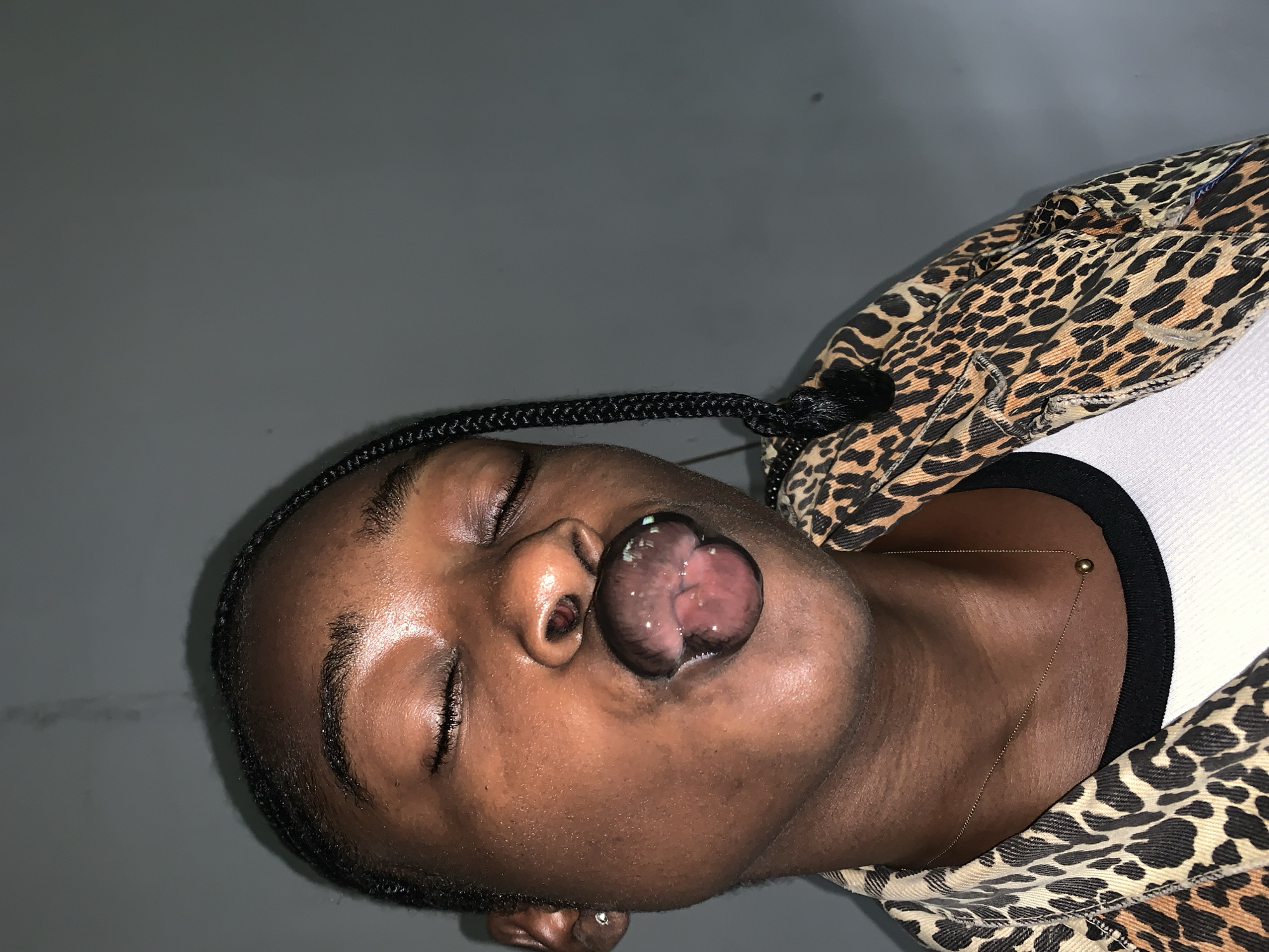 A medium shot shows a person of color with closed eyes and pursed lips, blowing a bubble. They have dark skin, braided hair that falls over their shoulder, and are wearing a leopard-print jacket over a white tank top with a black neckline. A delicate gold chain with a small sphere pendant rests on their neck. The background is a plain, gray wall. There is no indication of the specific location within Rafin Par, Nigeria.