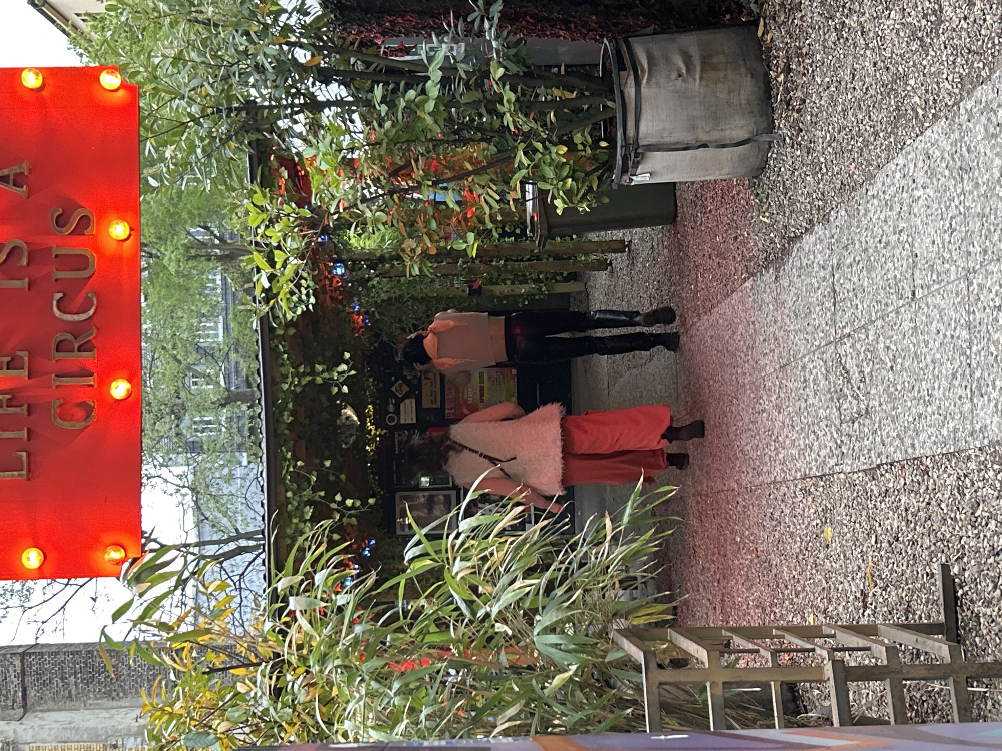 The image portrays an outdoor scene at an urban establishment in Berlin, Germany, possibly a cafe, bar, or entertainment venue. The setting is bright, suggesting daytime, though a prominent sign is illuminated.
On the left, a large red sign with glowing yellow bulbs clearly displays the text "LIFE IS A CIRCUS." The ground features a finely pebbled, light-colored surface in the immediate foreground, transitioning to a darker, coarser gravel path that leads into the background.
Two individuals are seen from behind, walking along the path away from the viewer. The person in the background wears a light-colored top, dark trousers, and dark boots. The individual closer to the foreground is dressed in a fluffy, light-colored jacket or vest over wide-legged red trousers, complemented by dark shoes.
The path is bordered by abundant greenery, including various potted plants and trees with both broad and narrow leaves, contributing to a lush ambiance. In the upper right, a gray, fabric-covered container or bag rests on the gravel. A wooden structure, possibly part of a railing or bench, is visible in the lower right, and reflections of a building facade can be glimpsed through the foliage in the background.