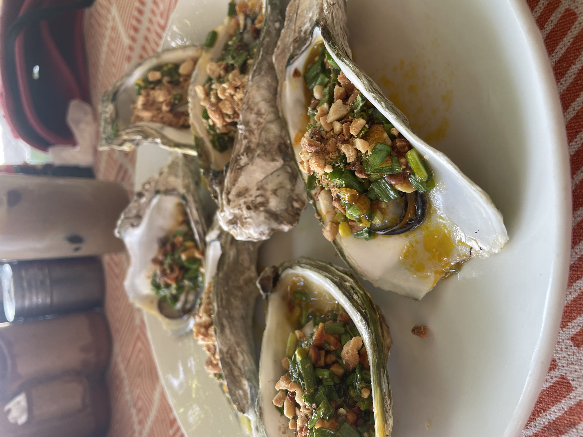 A plate of six grilled oysters topped with chopped green onions, chili, and crushed peanuts is presented on a white plate. The oysters are arranged on a table with an orange and white checkered tablecloth. In the background, a drink in a clear plastic cup and a red backpack are visible. The scene appears to be outdoors, possibly at a restaurant in Da Nang, Vietnam.