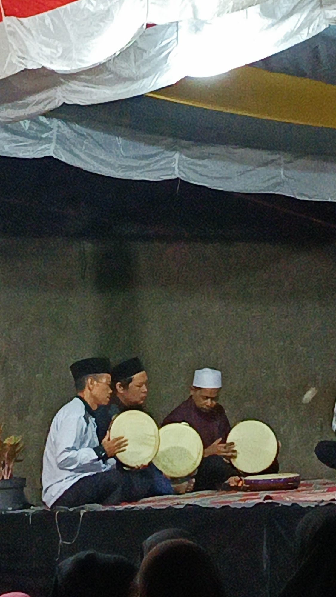 In Margajaya, Indonesia, three men are seated on a platform, engaged in playing percussion instruments. The man on the left, wearing a black cap and a light blue shirt, holds a tambourine. Next to him, a man in a black cap and dark shirt also holds a tambourine. The man on the right, dressed in a maroon shirt and a white cap, is playing a tambourine. Another tambourine rests on the platform in front of him. Behind the men, a textured, dark wall serves as the backdrop. The scene is illuminated by lighting, casting a warm glow. Above, a decorative canopy, likely made of fabric, is suspended, with sections of white and yellow fabric visible. The overall setting appears to be an indoor performance space, possibly a cultural event or religious gathering.
