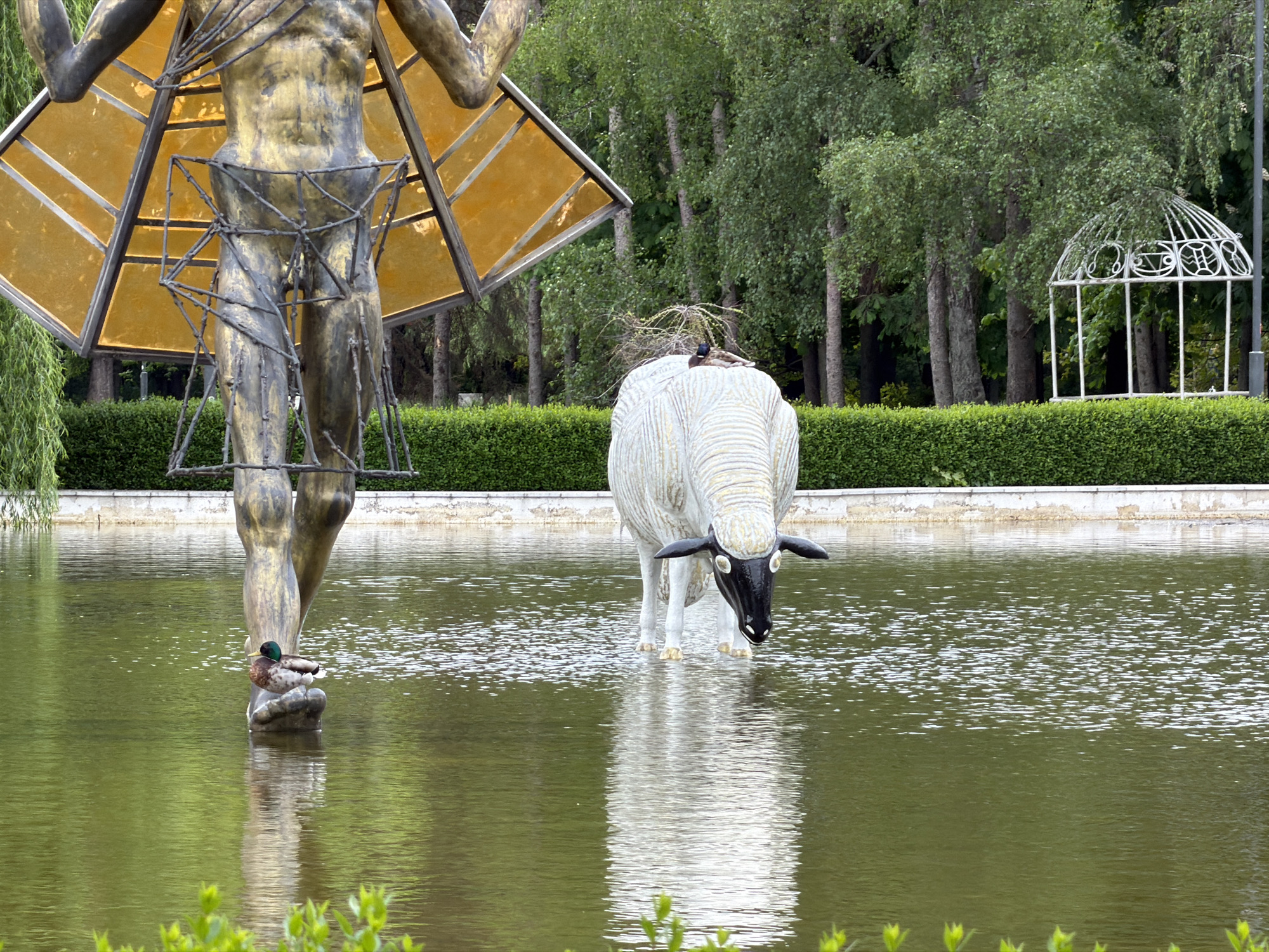 This photograph, taken in Sofia, Bulgaria, features a park pond setting. Dominating the foreground is a partially visible bronze sculpture of a man, clad in a geometric structure with golden panels. To the right, a white, stylized sculpture of a sheep with a black face stands in the water. A duck sits on the foot of the male sculpture. In the background, manicured hedges frame a small, decorative white gazebo. The pond's water surface reflects the sculptures and the surrounding greenery. Trees are visible behind the hedge.