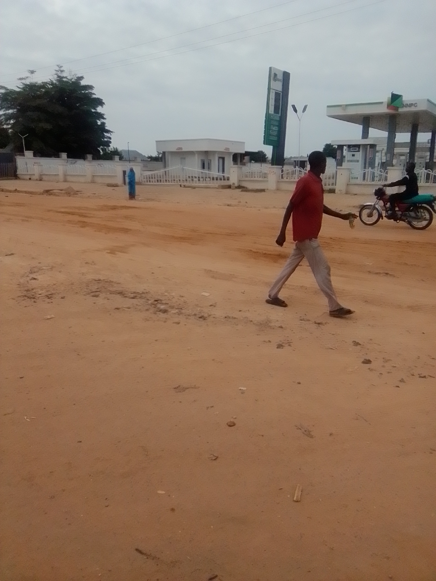 The image depicts a wide, unpaved dirt road in Gombe, Nigeria, under an overcast sky. In the foreground, a man in a red shirt and light-colored pants walks from left to right across the frame, carrying a light-colored object in his right hand. Behind him, a person rides a dark-colored motorcycle with a blue seat, also traveling from left to right.
In the middle ground, a white fence separates the road from a street lined with buildings. To the left, a large, dark green tree stands behind the fence. A blue figure, possibly a statue or a person in blue clothing, is visible near the fence. Further to the right, a white building with a prominent sign reading "NNPC" is visible, with a gas station canopy extending from it. A tall, green NNPC sign stands to the left of the gas station. The overall impression is one of a developing area with infrastructure, including a gas station, in a dusty, semi-urban environment.
