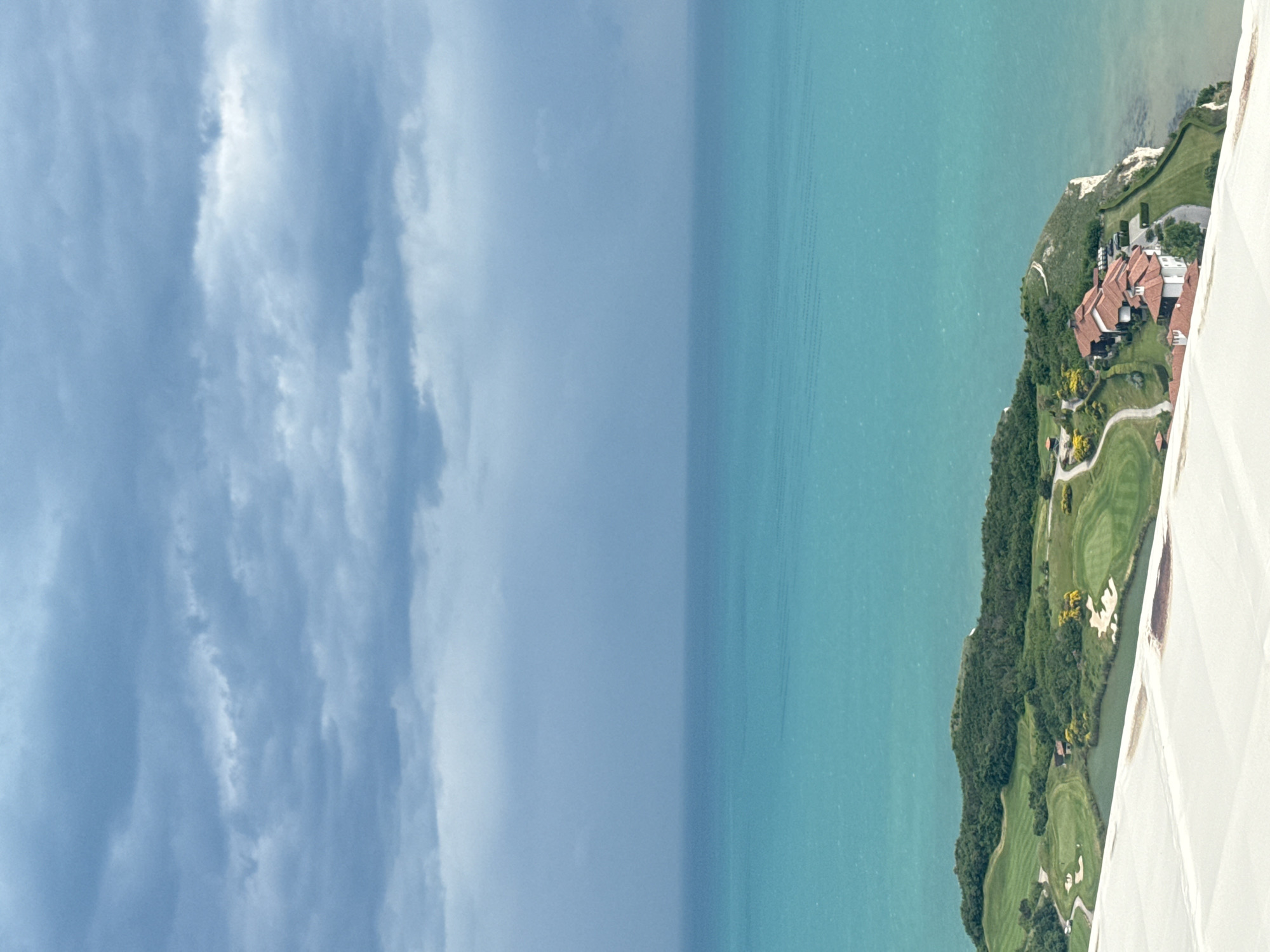 The image shows an elevated view of a coastal landscape. On the right, there's a landmass with lush greenery, featuring what seems to be a golf course and a residential area with red-tiled roofs. The land is bordered by a turquoise body of water that takes up the middle portion of the image. To the left, there's a cloudy sky, seemingly indicative of an overcast day. A portion of a white structure is visible in the lower right corner, from which the picture was likely taken. The image suggests a location near a scenic coastal area, possibly near the Black Sea. Given that Topola is located in Bulgaria, it's conceivable that the scene depicts a resort area near the Black Sea coast within Bulgarian territory.