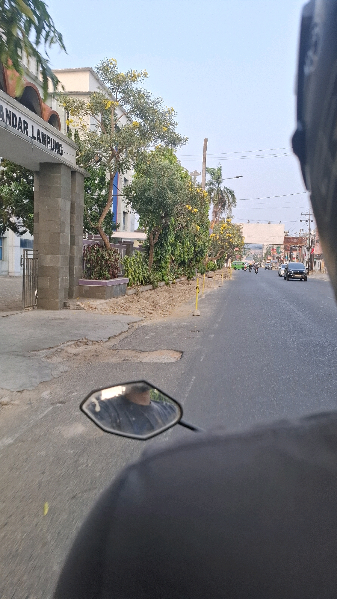 The image captures a street scene in Bandar Lampung, Indonesia, viewed from the perspective of someone riding a motorcycle. On the left, a building entrance with a gray stone archway and the text "BANDAR LAMPUNG" is visible. Adjacent to the entrance is a gate. Several trees, some with yellow flowers, line the sidewalk, which shows signs of disrepair with cracked pavement and exposed dirt. Yellow bollards are placed along the edge of the road. The asphalt road itself has several potholes. In the distance, traffic can be seen, including cars and motorcycles, as well as a green bus. Buildings and utility poles are also present along the street. A motorcycle's rearview mirror in the foreground reflects the rider and the sky. The sky is clear and light blue, suggesting it is daytime.