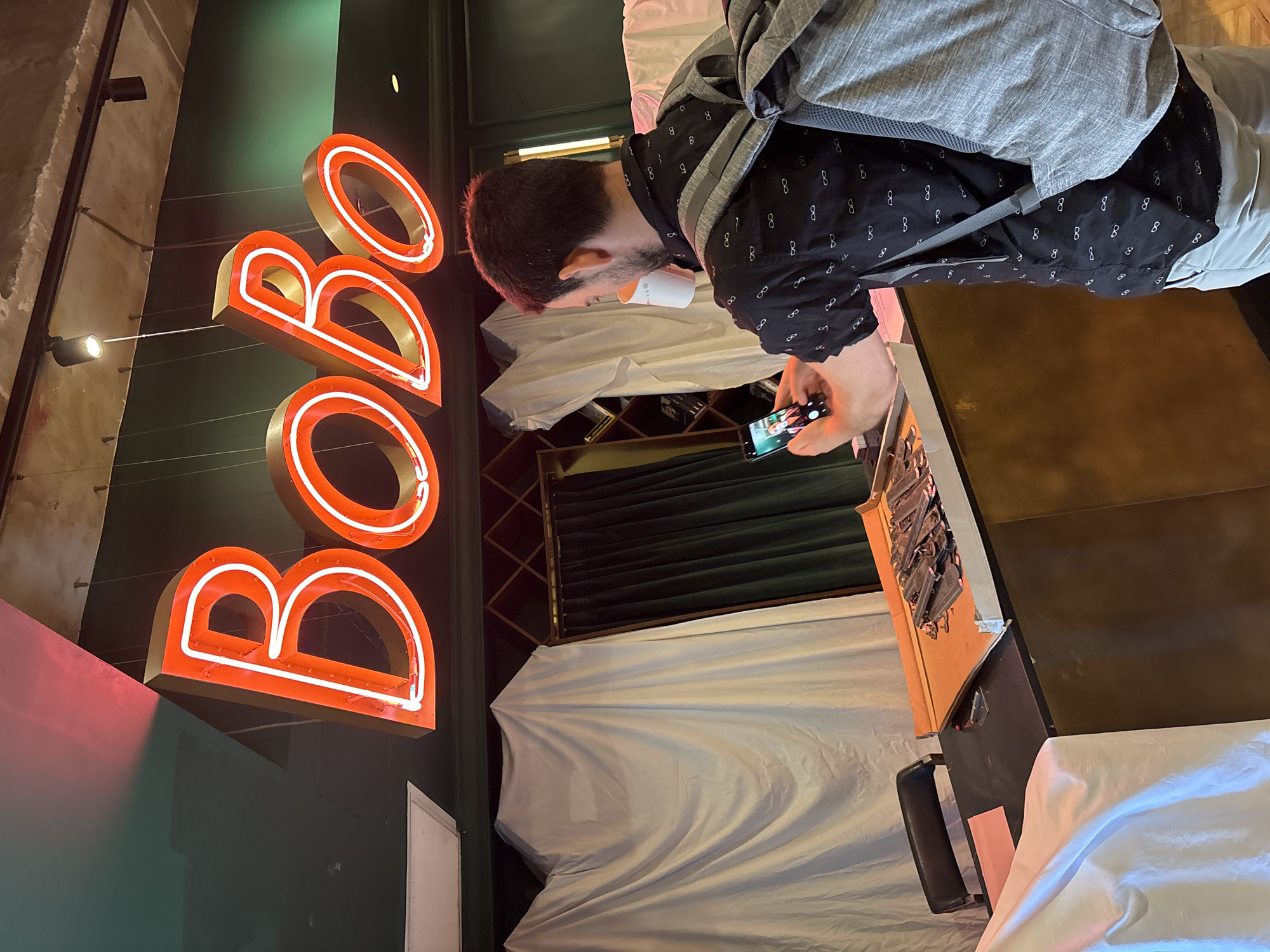 An indoor scene in Tel-Aviv, Israel, features a large, upright neon sign glowing in orange-red, prominently spelling "BOBO" against a dark green wall. In the foreground, a man with dark hair and a beard, wearing a black collared shirt patterned with white infinity symbols and a grey backpack, stands focused on his smartphone, which he holds with both hands. The phone's screen displays an image. A white cup is visible near his face, suggesting he may be drinking from it.
Various surfaces below the neon sign and around the man are covered with white drapes or sheets, possibly indicating the establishment is closed, undergoing renovation, or being prepared for an event. A dark, possibly wooden, counter or table runs along the right side of the frame, partially covered. Small spotlights are mounted on a lighter-colored wall to the left of the neon sign. The overall lighting is dim, with the bright neon sign providing a significant light source, suggesting an evening or night setting for this commercial space, likely a cafe, bar, or restaurant.
