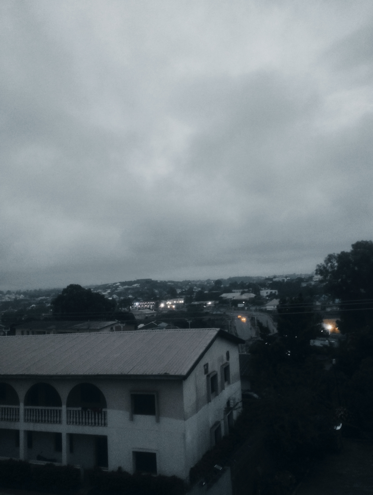 This image depicts an overcast sky over a residential area in Jos, Nigeria. In the foreground, a two-story building with a sloped metal roof and arched balconies is visible. The building's facade is painted in light and dark shades, and some windows are dark openings. The middle ground shows a landscape of houses, some with illuminated windows, suggesting evening or nighttime. Trees and vegetation are scattered throughout the scene, some appearing dense. The background consists of a hazy sky with clouds and a distant cityscape or more houses. The overall atmosphere is subdued due to the cloudy weather and low light conditions.