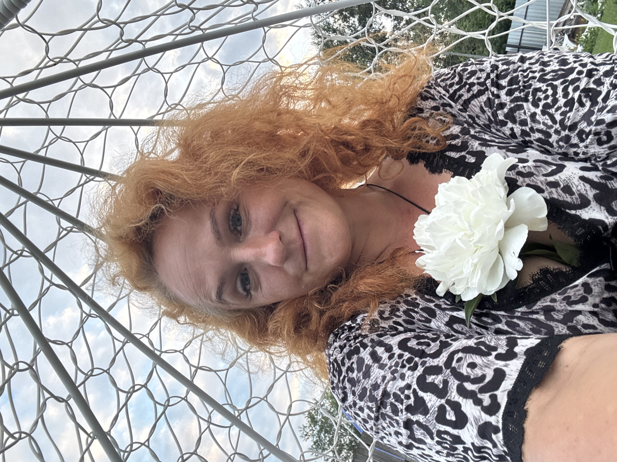 In the city of Chaikovsky, Russia, a woman with voluminous, curly red hair is pictured in a selfie. She is seated within a white, geodesic-like structure, possibly a garden swing or hammock, constructed from interconnected white metal rods and netting. The background shows a sky with scattered clouds and the tops of trees. The woman is wearing a black and white leopard-print top with black lace trim. A large, white peony flower is pinned to the front of her garment, near her chest. Her expression is a soft, closed-mouth smile, and she appears to be looking directly at the camera. The overall impression is a casual, outdoor portrait.