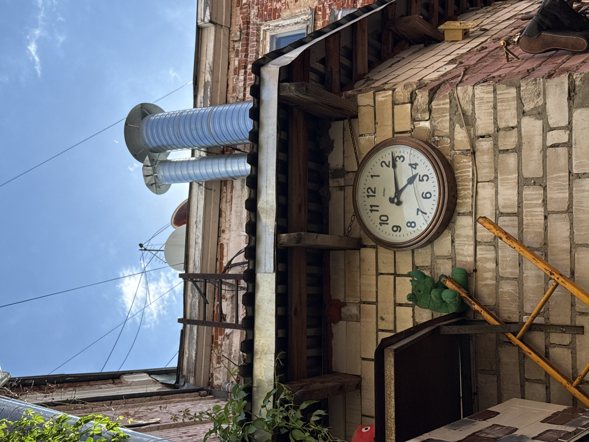 The image depicts a close-up view of the exterior wall of a brick building, likely in an urban setting. A large, circular analog clock with Roman numerals is prominently mounted on the lighter colored brickwork. The clock hands indicate a time between 4:15 and 4:20. To the left of the clock, a green stuffed toy, resembling a rhinoceros, is perched on a yellow ladder. The ladder is leaning against the wall. Above the clock and ladder, the roofline of the building is visible, featuring a corrugated metal roof and exposed wooden beams. Two large, silver corrugated metal ventilation pipes extend upwards from the roof, against a bright blue sky with scattered clouds. The brickwork shows signs of wear and tear, with some cracks and staining. The overall scene suggests a somewhat weathered and possibly bohemian or artistic urban environment. The context of Nizhny Novgorod, Russia is not visually apparent from the image itself.