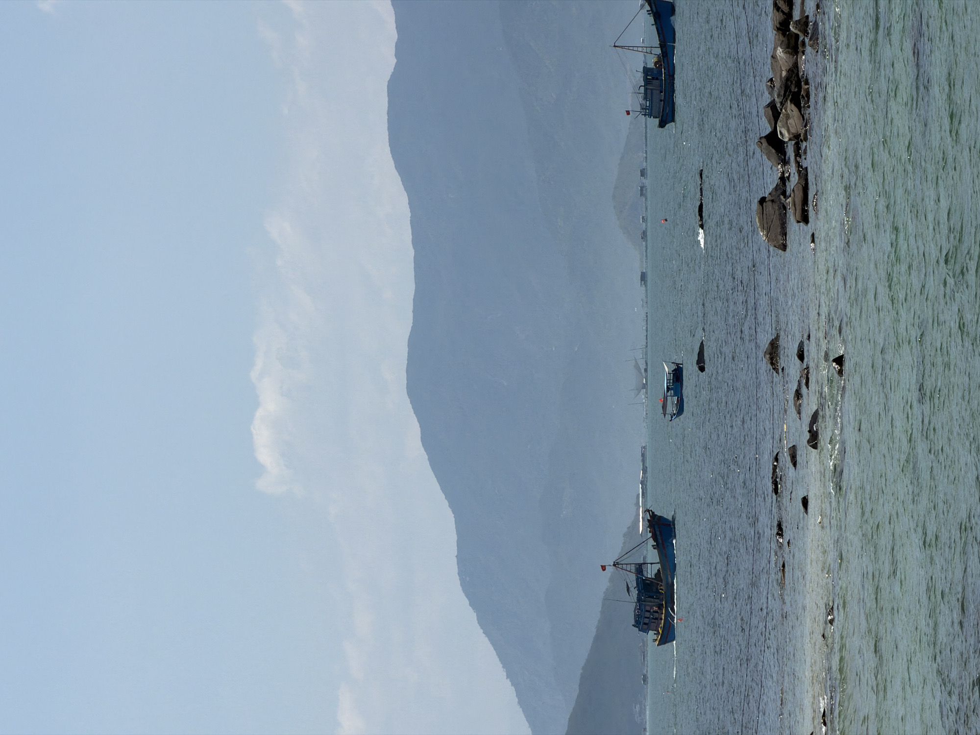 This image depicts a coastal scene in Vĩnh Lương, Vietnam, under a bright, hazy sky with scattered white clouds. The horizon features a series of distant, layered mountains, rendered in muted blue-grey tones due to atmospheric perspective, extending across the width of the frame. The water in the midground is a deep greenish-blue, showing slight ripples and patches of sunlight reflection.
Several boats are visible on the water. In the left-midground, a prominent blue and yellow fishing boat with a red flag affixed to its mast moves towards the right, leaving a discernible wake. Its structure includes an elevated platform and various rigging. To the right of the center, a smaller, solid blue boat with a white canopy is positioned. Further to the right, another large blue fishing boat, similar in design to the one on the left but appearing slightly larger, is visible, also featuring a red flag and an elevated structure. In the far distance, numerous small, dark structures, possibly floating houses or smaller boats, are scattered across the water near the base of the mountains. A red buoy is also present in the midground water between the central small boat and the rightmost large boat.
The foreground water is clearer and shallower, revealing numerous dark grey rocks of varying sizes partially submerged or breaking the surface. The water color in the foreground is a lighter, more translucent green. No individuals are explicitly visible in the frame, though the presence of the boats implies human activity.