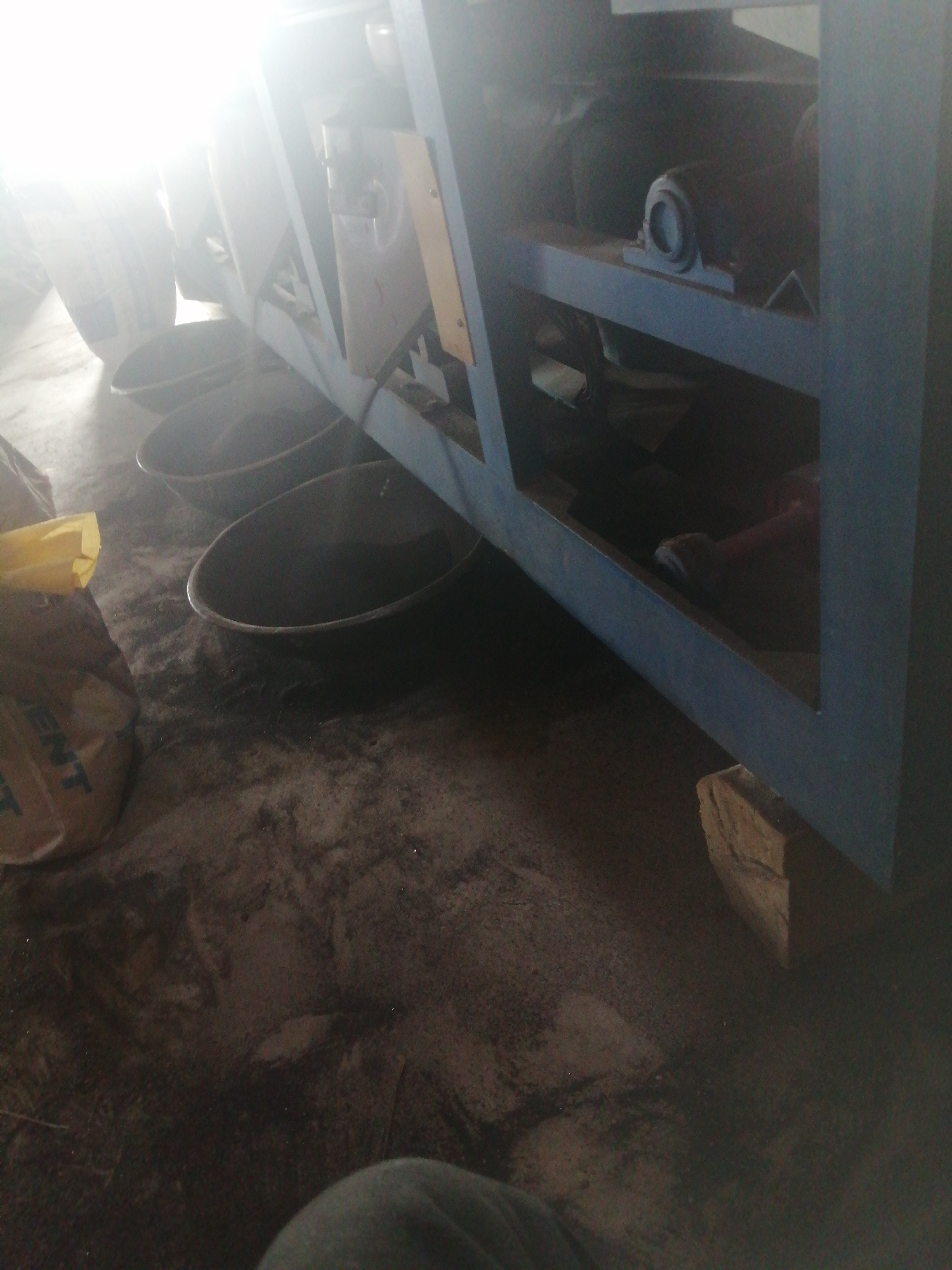 The image shows a ground-level view of an industrial or workshop setting, likely in Jos, Nigeria, characterized by a concrete floor covered in dust and debris. In the foreground, there is a brown bag with yellow lettering, possibly containing materials like cement or construction aggregate, a common sight in Jos. Behind the bag, a large, dark, basin-like object is visible, suggestive of a mixing tub or a collection receptacle. Further back, other similar basins are partially obscured. The background is dominated by a blue metal structure, part of a larger machine or frame, with shelving and some mechanical components, including what appears to be a bearing housing. The lighting is harsh and bright from the upper left, creating significant glare and obscuring details in that area, possibly indicating the presence of strong artificial or natural light sources. The overall impression is one of an active but somewhat unkempt workspace.