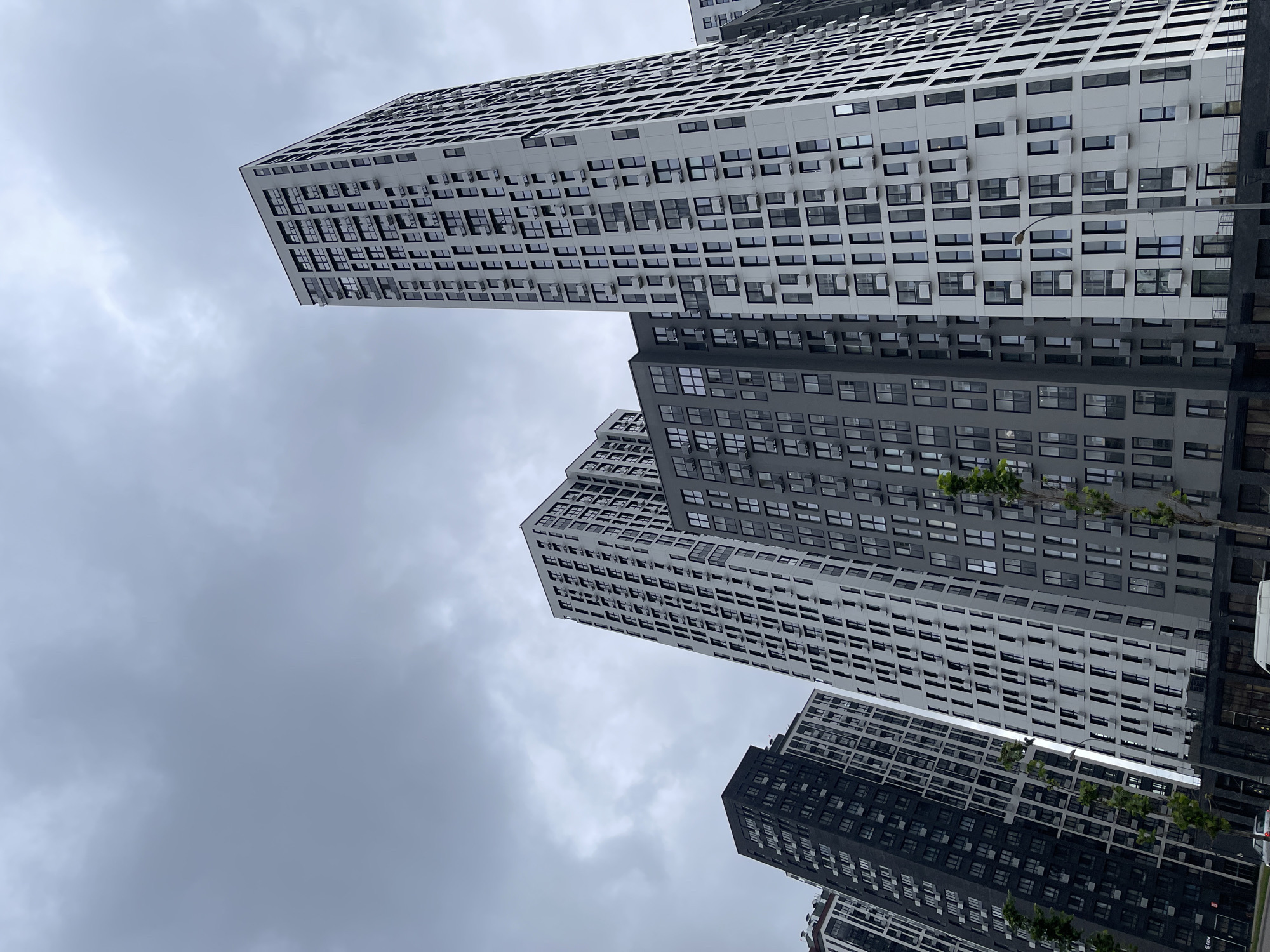 This is an outdoor, eye-level, medium shot of a cluster of modern, high-rise apartment buildings in Moscow, Russia. The buildings are tall and narrow, with many windows and balconies. They appear to be made of concrete and glass. The sky is overcast and grey, suggesting a cloudy day. The buildings are of various heights and colors, ranging from light grey to dark grey and black. Some buildings have a pattern of dark vertical lines. In the foreground, there is a street with a few cars and a white van. There are also some trees lining the street. The scene appears to be a residential area of a modern city.