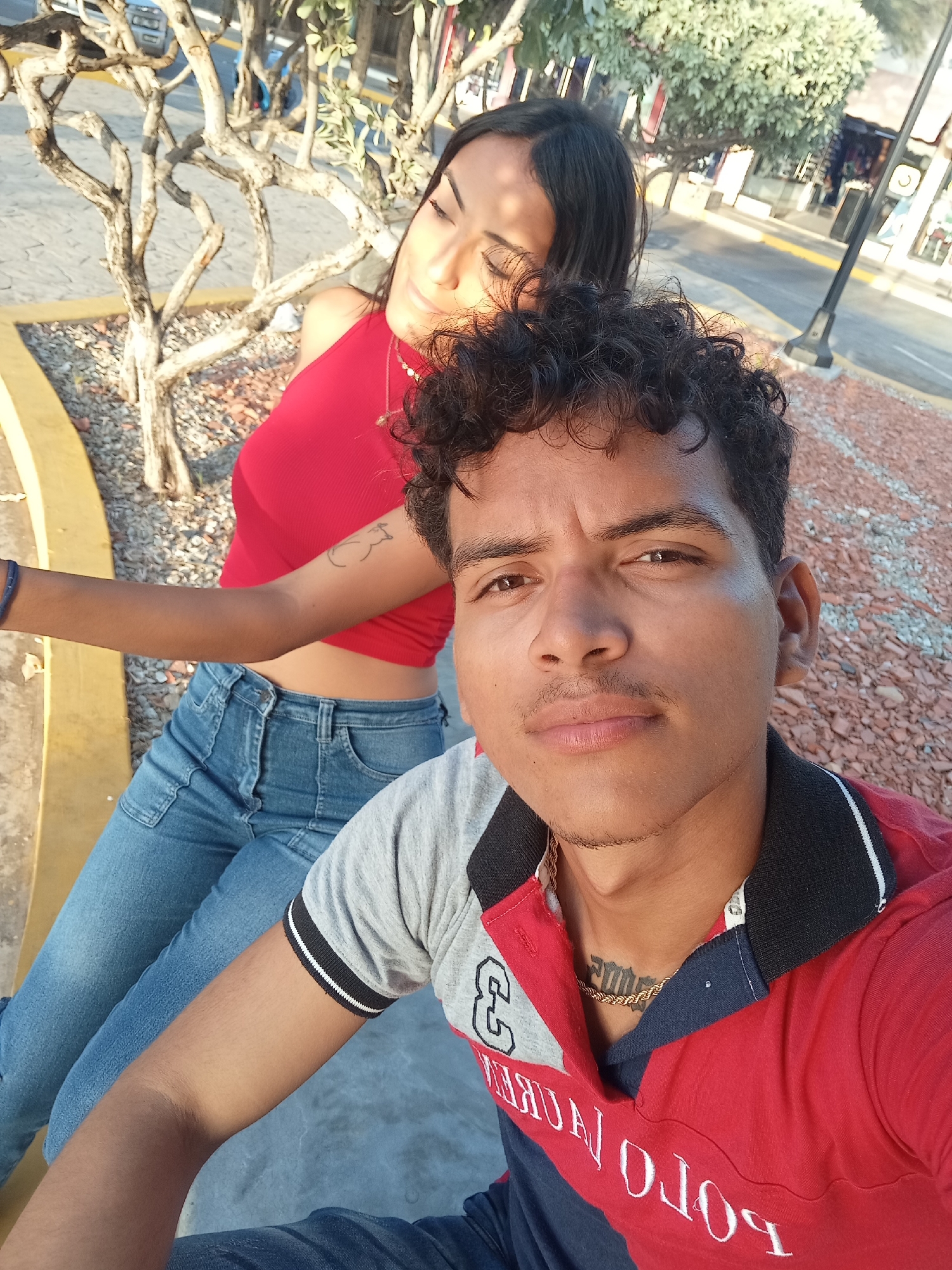 The image shows a selfie of a man and woman sitting outdoors. The man is in the foreground, wearing a red, gray, and navy blue polo shirt with a gold chain and a tattoo on his neck. The woman is sitting behind him, wearing a red halter top and blue jeans. She has a tattoo on her arm. They are sitting on a concrete bench next to a tree. The background shows a paved area with buildings and streetlights, indicative of a public space or plaza. The location is likely in the city of Juan Griego, Venezuela, based on the user's prompt. The scene appears to be a casual moment captured during the day.