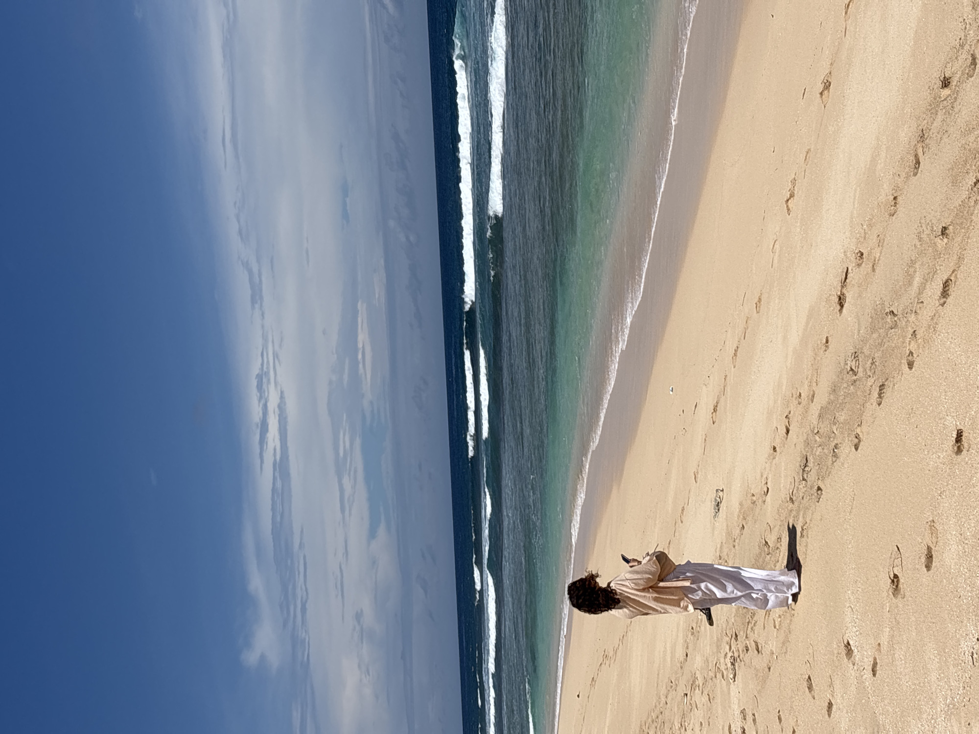 The image shows a beach scene in Pecatu, Indonesia. The sky is partially clear with blue sky and some white clouds. The ocean has waves breaking near the shore. A woman stands on the sandy beach. She has dark curly hair and wears a light tan shirt or jacket and white pants. She is holding a phone and looking down at it. There are footprints in the sand around her.