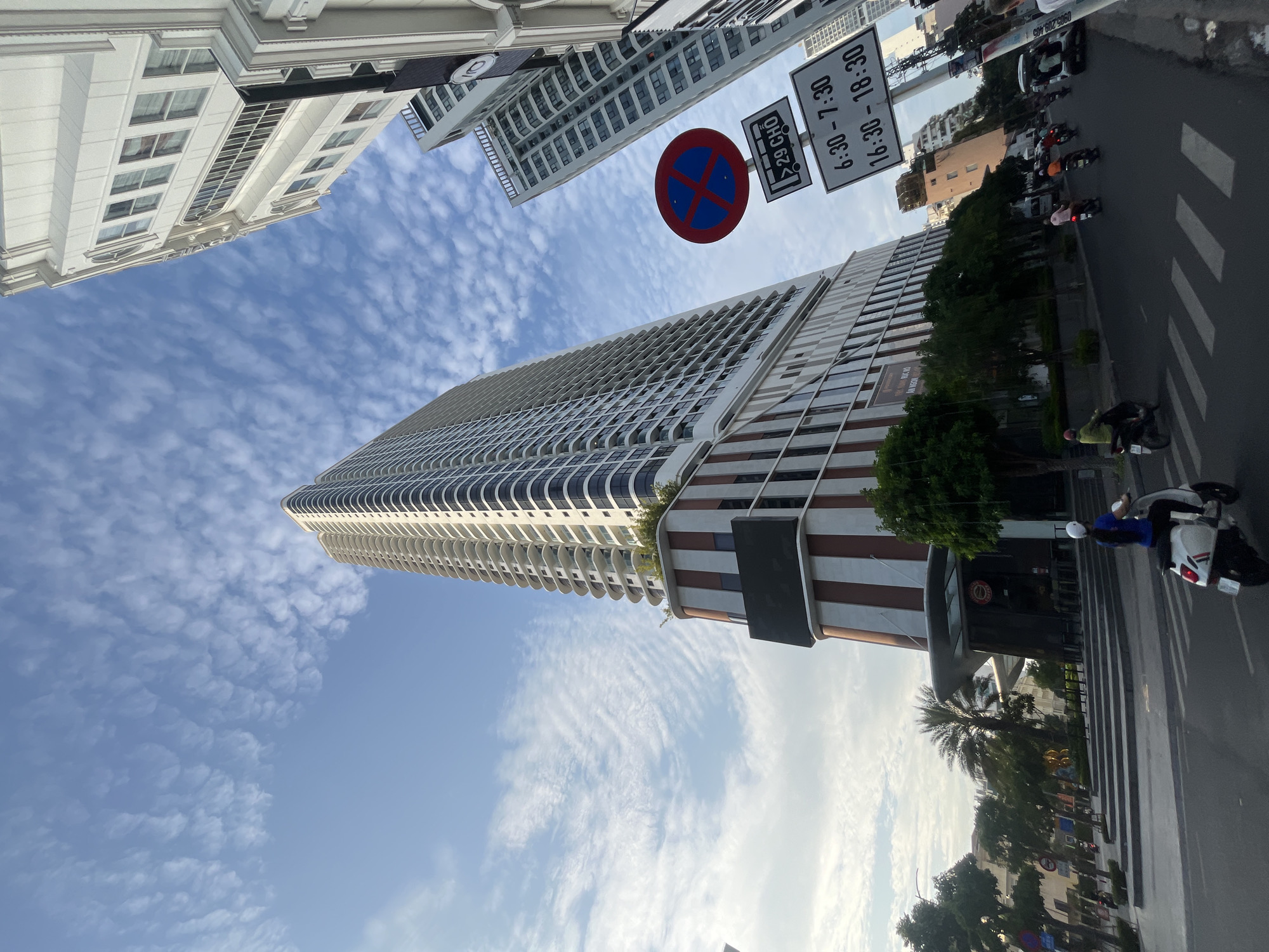 A low-angle shot captures a modern skyscraper dominating the frame, its facade a repeating pattern of balconies and dark-tinted windows. The building is situated in an urban environment, with other high-rise buildings visible in the background. A street runs in front of the skyscraper, with a few motorbikes navigating the road. A woman on a white motorbike, wearing a white helmet and blue top, is the most prominent human figure, riding away from the camera. Traffic signs, including a no-parking symbol and time restrictions, are mounted on a pole to the right of the skyscraper. The sky above is a bright blue, dotted with wispy, white clouds, suggesting fair weather. Trees and some shrubbery are present on the sidewalk and in front of the buildings, adding a touch of greenery to the urban landscape. The scene appears to be in Nha Trang, Vietnam, a coastal city known for its modern architecture and busy street life.