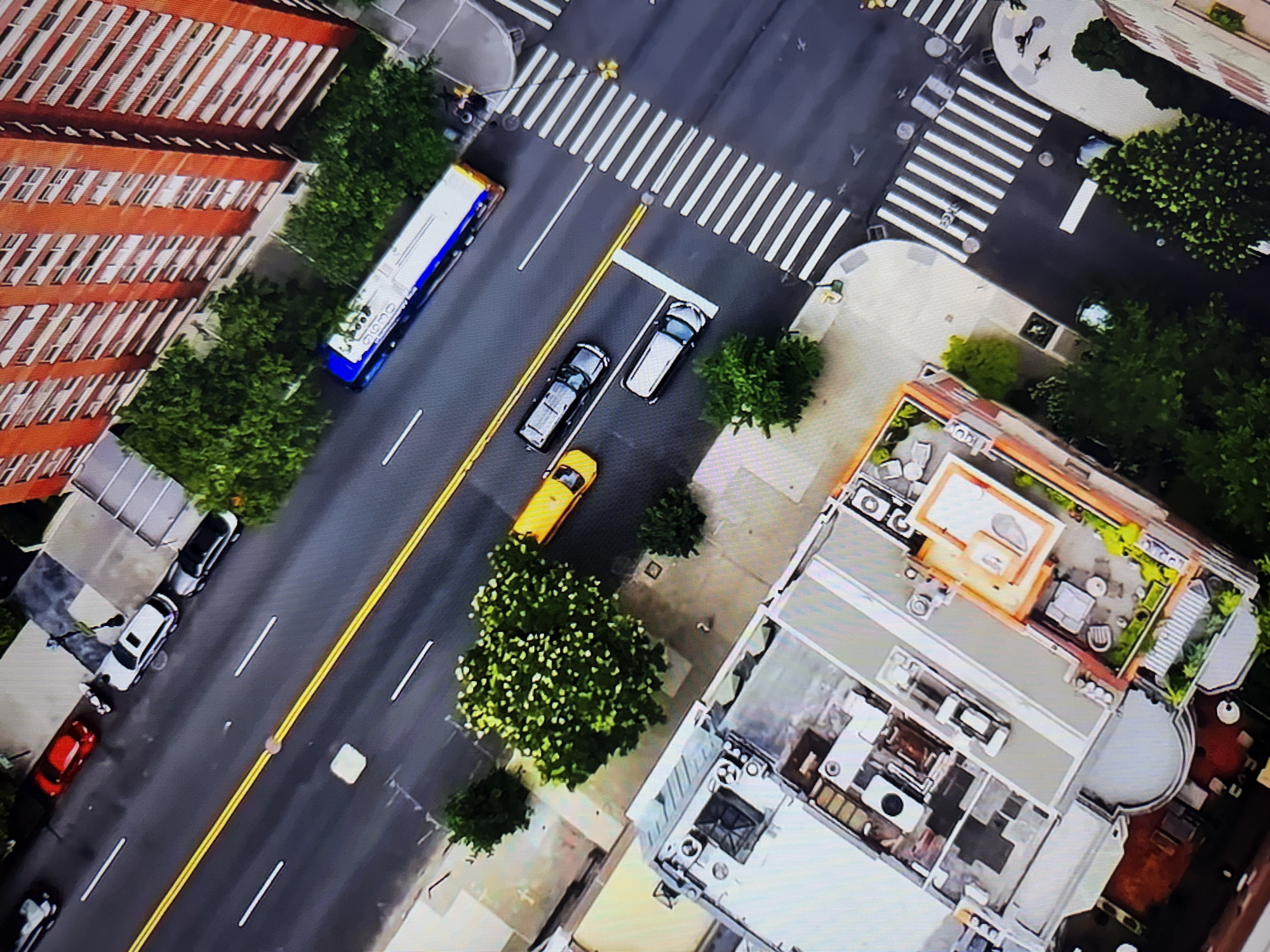 The image is an aerial view of a city street scene. In the foreground, there are several buildings, some with rooftop terraces. One building has outdoor furniture and greenery on its terrace. Further up the street, there are trees lining the sidewalk.
The street itself has yellow double lines dividing the lanes. Several vehicles are visible: a blue and white bus, a yellow taxi, and at least three other cars. The cars are parked and driving on the road. There are also pedestrian crosswalks visible at an intersection.
In the background, a tall, brick building with many windows is prominent. The overall impression is a busy urban environment with a mix of residential and commercial buildings, traffic, and green spaces.
Without specific visual cues or landmarks that are unique to Burgas, Bulgaria, it is impossible to confirm the location.