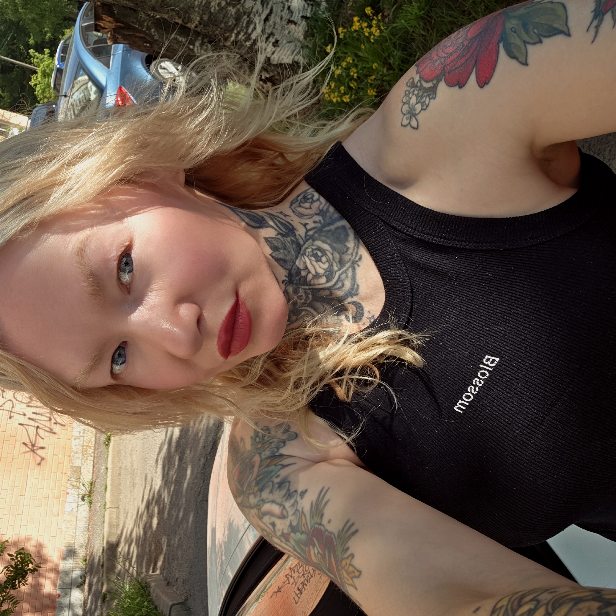 A woman with light blonde hair, blue eyes, and red lipstick is the central subject of the image. She is wearing a black tank top with the word "Blossom" printed on it. Visible tattoos adorn her neck and arms. The backdrop includes a street scene, suggesting an urban environment. A blue car is partially visible in the background. Brick buildings and street markings also indicate a city. The presence of these elements, along with the yellow flowers, suggest that the image was taken in Saint Petersburg, Russia.