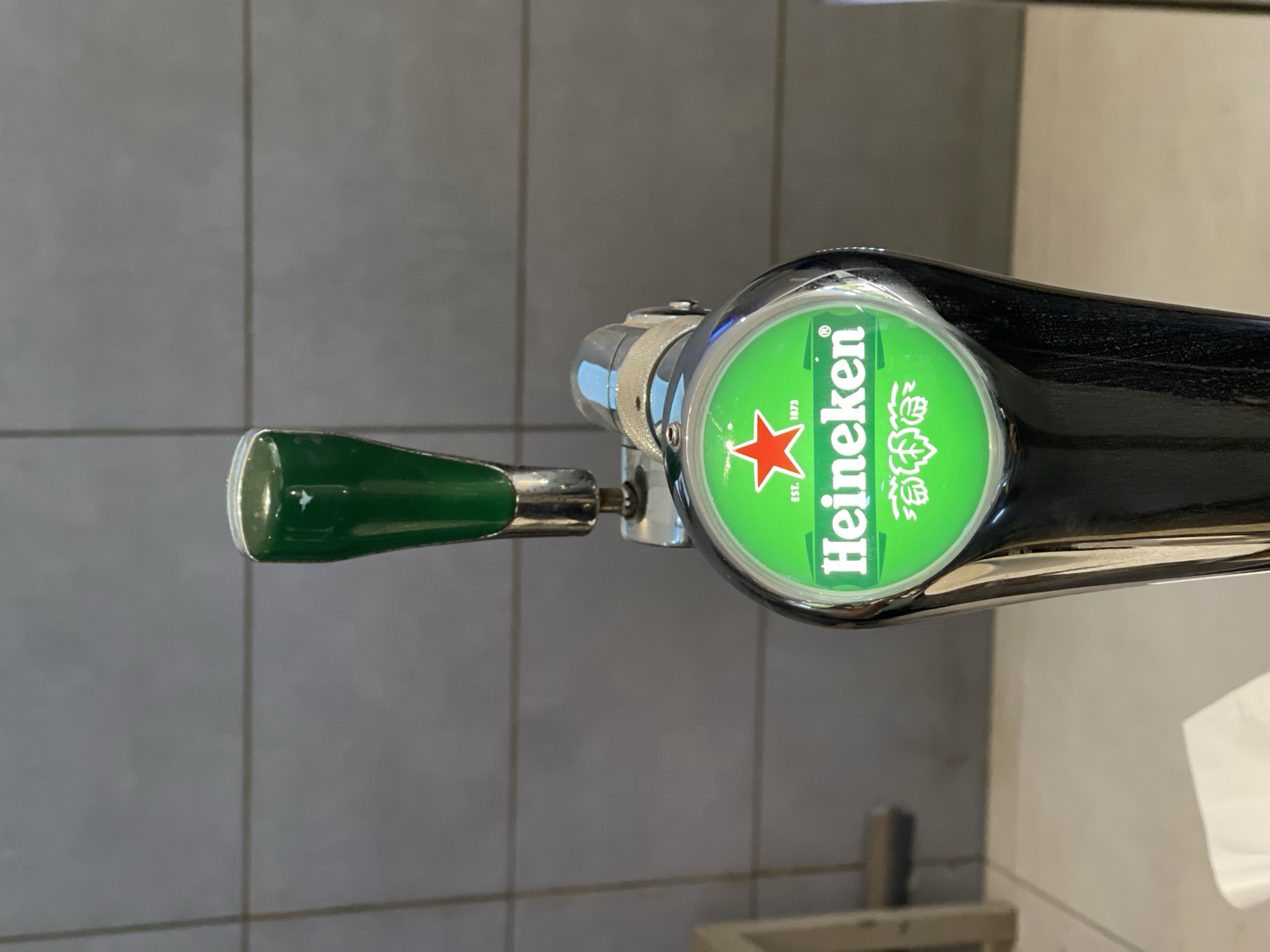 The media file captures a close-up view of a Heineken beer tap, oriented horizontally due to the image's rotation. This draft beer dispenser is likely part of a bar or restaurant setting in Abuja, Nigeria.
The tap features a polished chrome body and a distinct green handle, which points to the left in the current orientation. At the top of the tap, a prominent circular finial displays the iconic Heineken logo on a vibrant green background. The logo includes a bright red five-pointed star, the brand name "Heineken" in white block letters, and the establishment date "Est. 1873". To the right of the brand name, a stylized white hop design is visible. The tap's main body below the finial appears dark and glossy.
In the background, a tiled wall with light grey, rectangular tiles is visible. The horizontal and vertical grout lines suggest a neat, possibly commercial, interior space. Towards the lower right of the frame, a lighter, off-white or beige surface, potentially a wall or counter, and a crumpled white object (possibly a napkin or tissue) are faintly discernible. The overall scene indicates an indoor environment with ambient lighting, reflected by the shiny surfaces of the tap. No people are visible, and no specific event or activity is underway, though the presence of the tap implies a beverage service area.