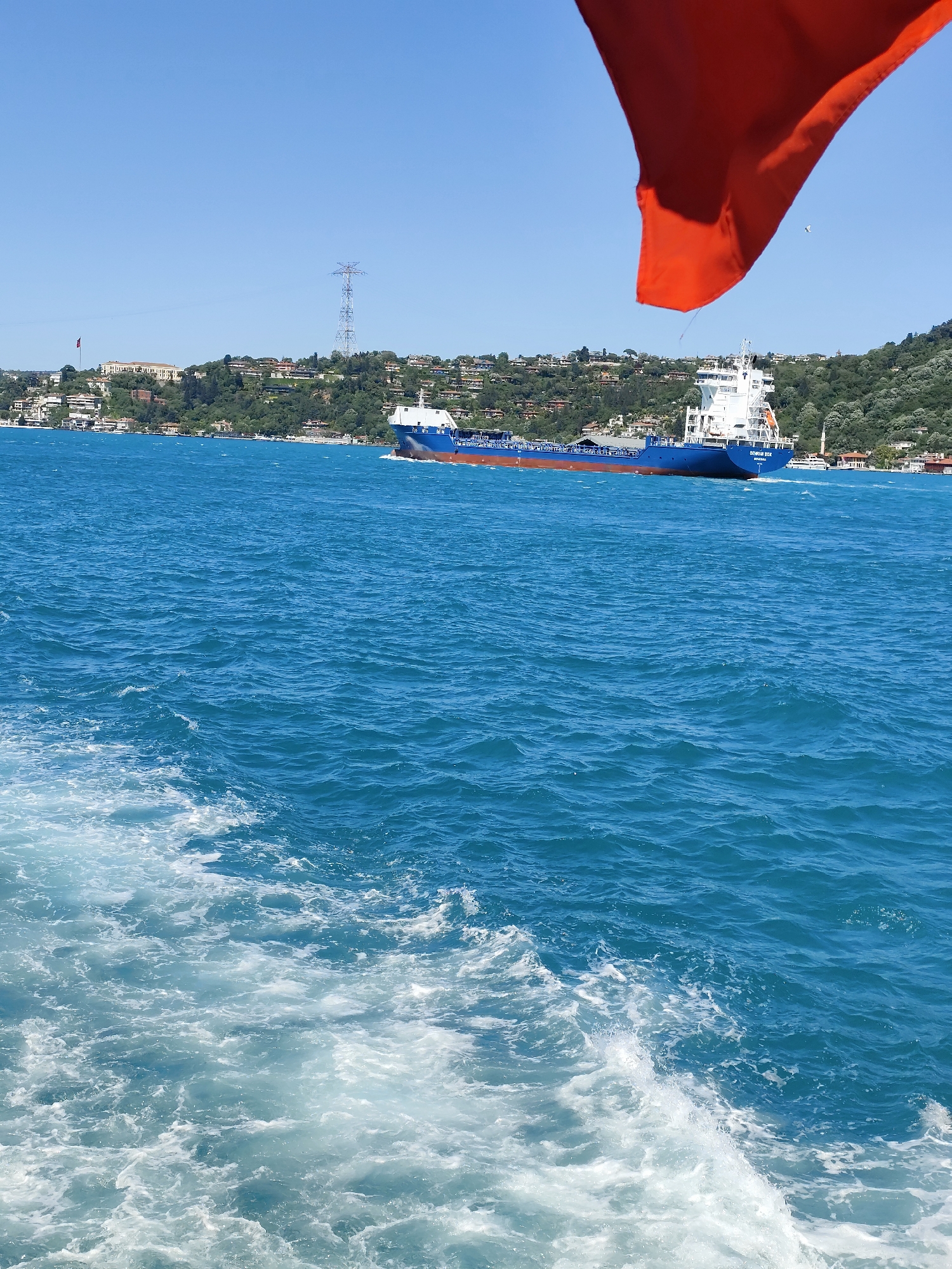 The image captures a view of the Bosphorus strait, likely taken from a moving vessel in Beşiktaş, Turkey. The foreground is dominated by the turquoise water, churned into frothy white waves. In the middle ground, a large cargo ship with a blue hull and white superstructure moves across the water. The background features a coastline with lush green vegetation, dotted with buildings. An electricity pylon is also visible. A large orange flag is visible in the top right corner.