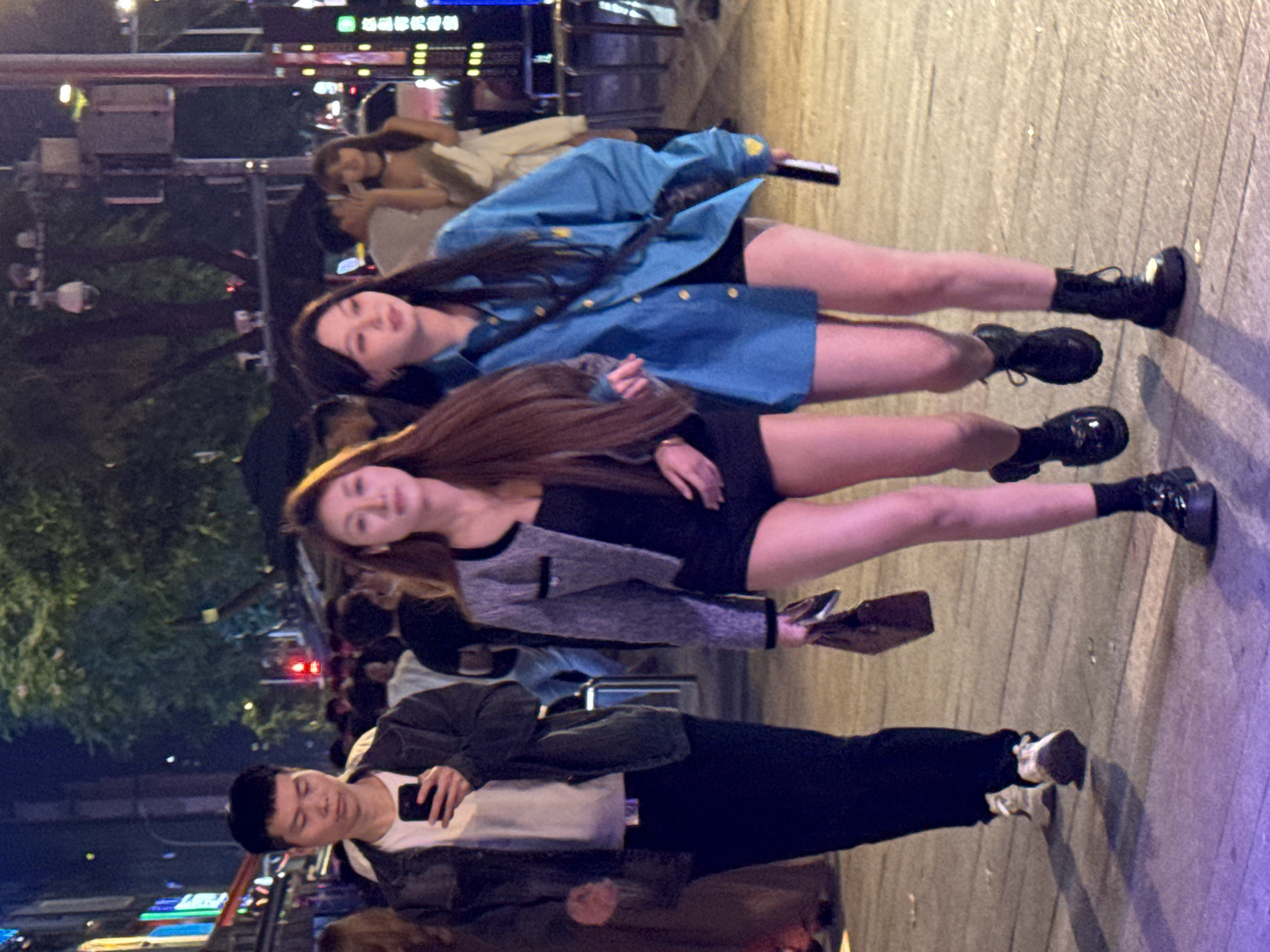 The image shows a group of people on a city street in Liangjiang, China, at night. Two young women are in the foreground, both wearing short dresses and boots. One woman is wearing a blue denim jacket, and the other is wearing a gray blazer. A man stands in front of them, looking at his phone. He is wearing a dark jacket, white shirt, and dark pants. Other people are walking in the background, and there are buildings and streetlights visible.