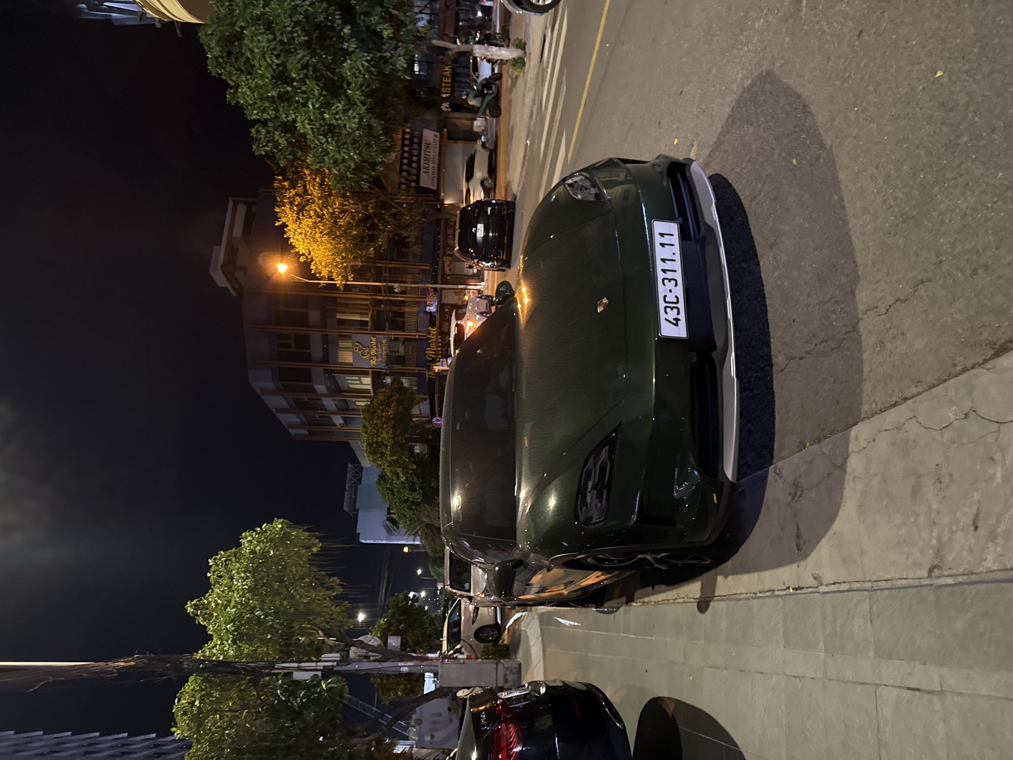 An exterior, eye-level, wide shot captures a dark green Porsche Taycan parked at the side of a street at night in Đà Nẵng, Vietnam. The vehicle is facing slightly to the right, with its front bumper and license plate, "43C-311.11," clearly visible. The street is paved, with a sidewalk to the left of the car. The sidewalk is made of square concrete tiles and appears slightly weathered. Beyond the sidewalk, a line of trees with lush green foliage is visible, their branches illuminated by streetlights. In the background, several buildings line the street, featuring commercial signage. One sign reads "Thai Market Restaurant," and another says "AKIMITSU JAPANESE RESTAURANT." The sky is dark, indicating nighttime. Other vehicles, including a black SUV and a white sedan, are parked or driving further down the street. A person on a scooter is also visible in the distance. The scene is illuminated by a combination of streetlights and ambient lighting from the buildings.