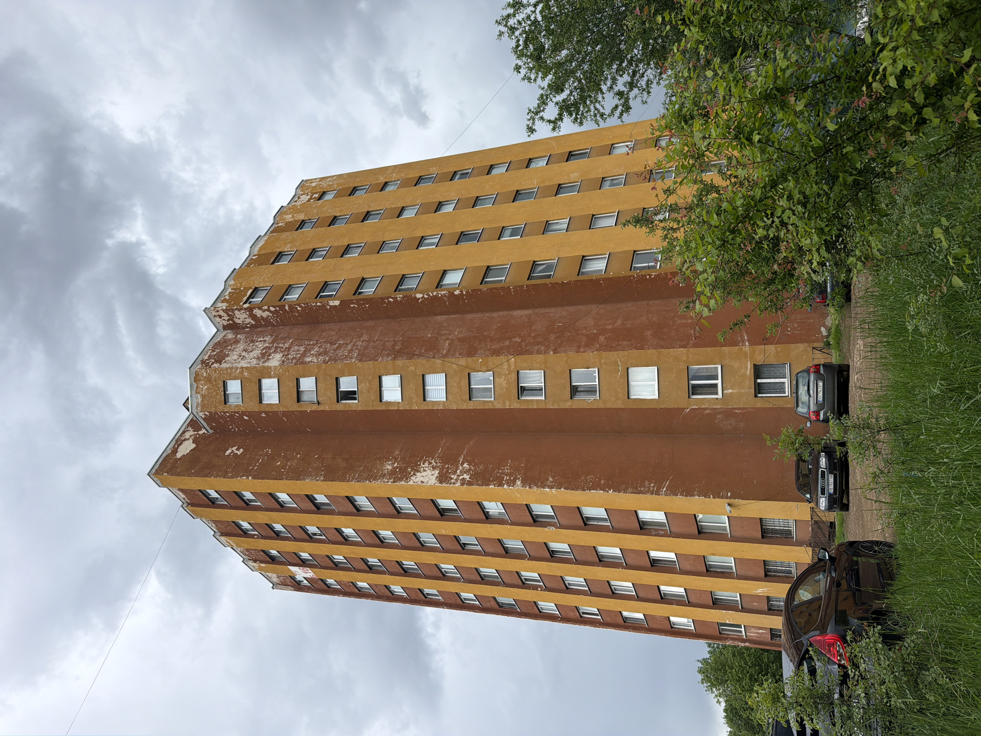 A tall, ochre-colored, rectangular building with a jagged top dominates the frame. Brown patches are visible near the middle of the building. The building features a uniform grid of windows along its facade. Overcast, grey skies fill the background. Trees and grass line the side of the frame. Several cars are parked near the building. The image shows a building in Sofia, Bulgaria.