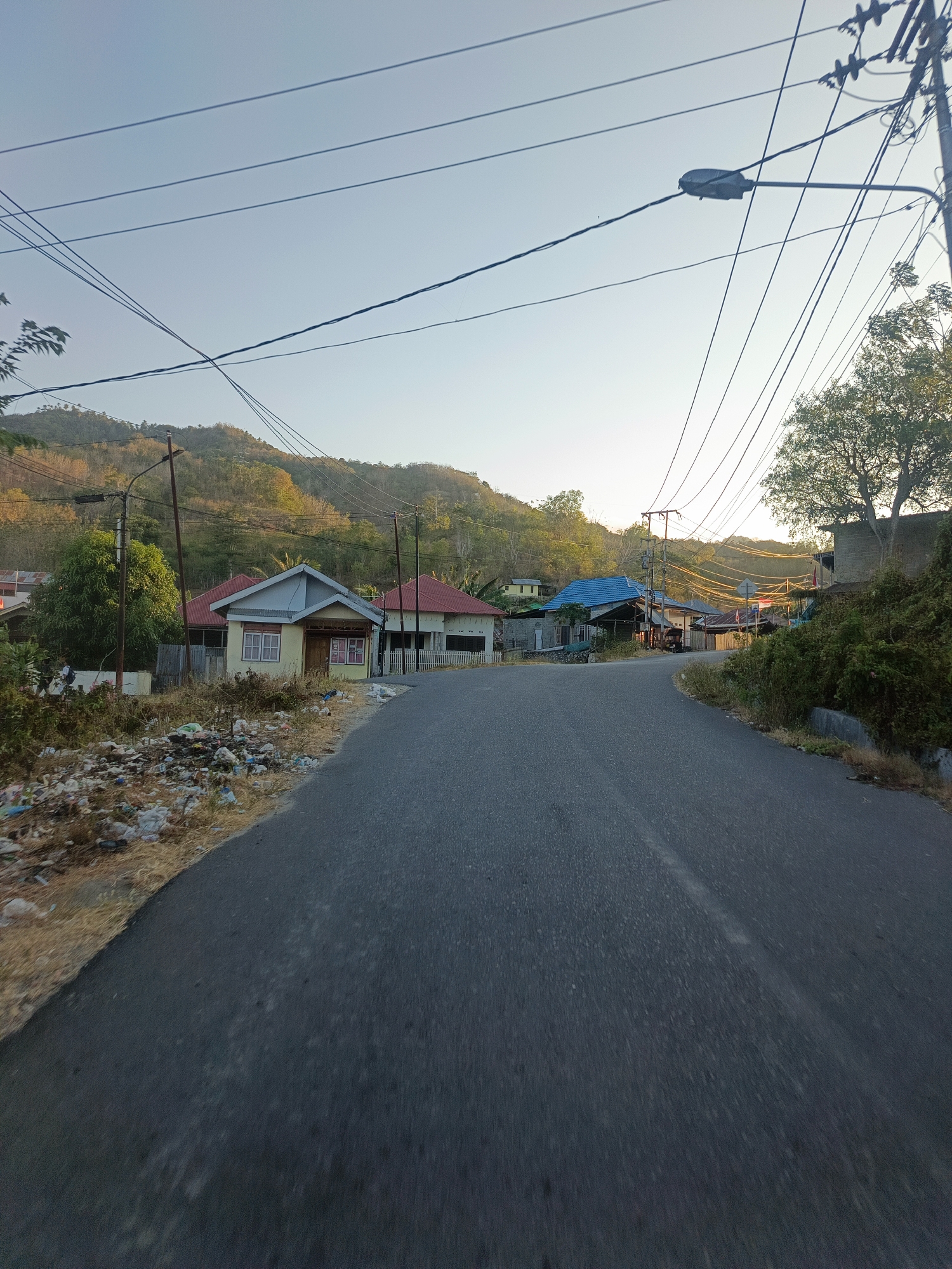 The image depicts a rural road in the city of Gorontalo, Indonesia, on a clear, bright day. The road curves gently to the right, with houses and vegetation lining its left side. The houses are modest, single-story structures with red and blue roofs. The immediate left of the road is cluttered with debris, suggesting a lack of waste management. Overhead, a network of power lines stretches across the sky, supported by wooden poles. A streetlamp is also visible. In the background, a tree-covered hill rises, bathed in sunlight. The overall impression is one of a developing area, with the natural landscape juxtaposed with human settlement and infrastructure.