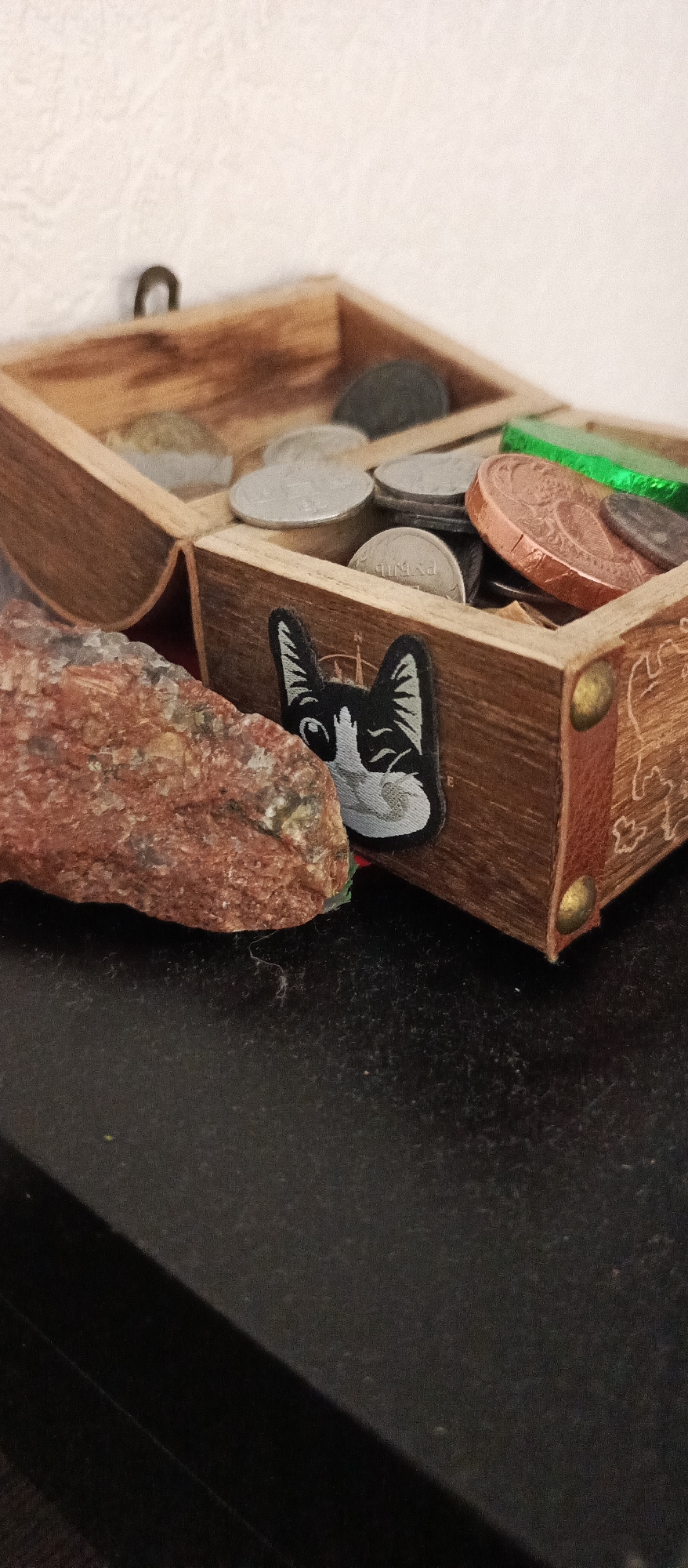 A close-up, eye-level shot shows a wooden box filled with various coins. The box is positioned on a dark, textured surface. To the left of the box, a reddish-brown, rough-textured rock is visible. On the front of the box, a black and white patch of a winking cat's face is affixed. The background is a textured, off-white surface. The scene is lit from the front, casting a shadow to the right.