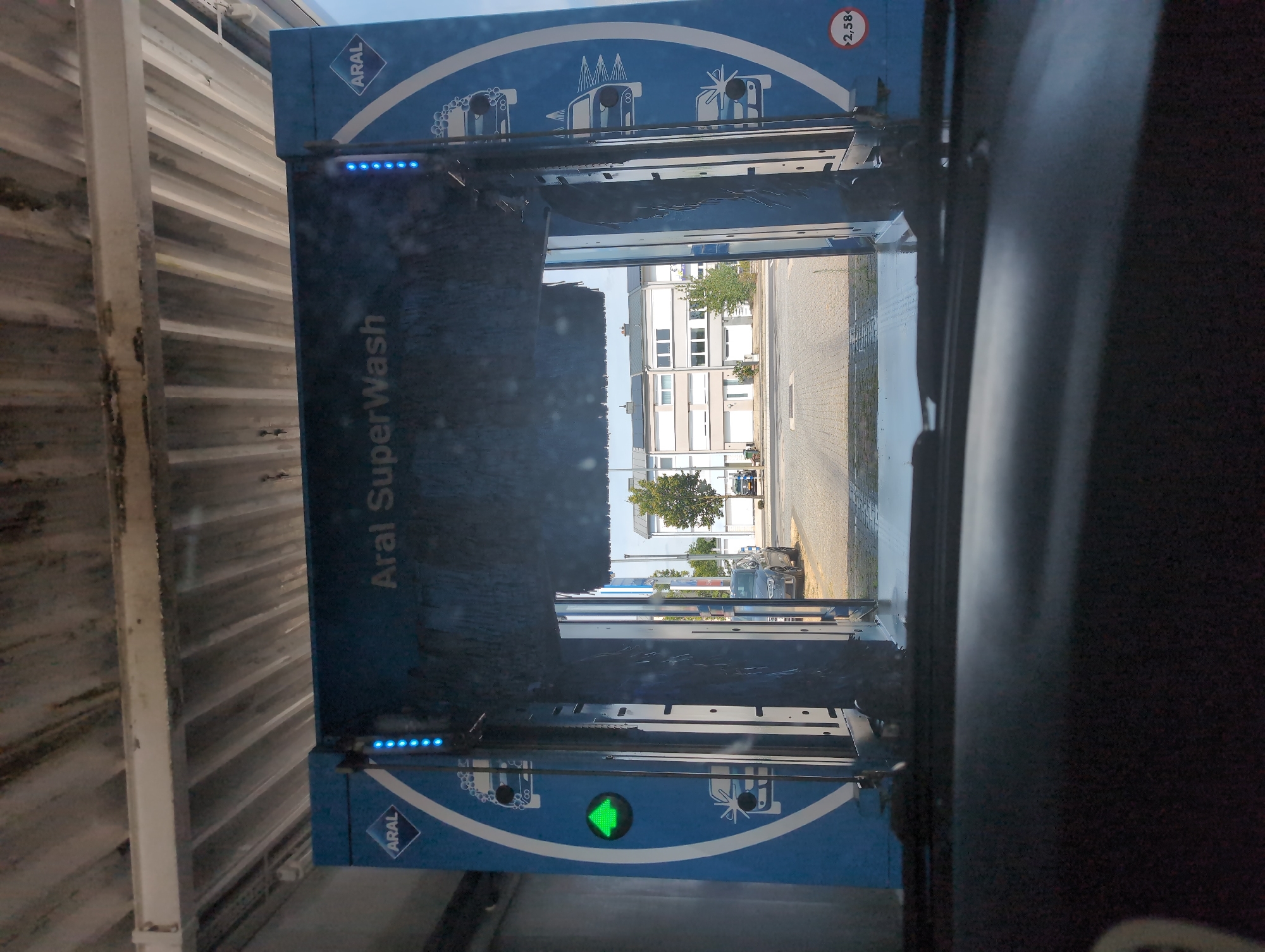 The image captures the interior of a car driving into an Aral SuperWash car wash. The car wash machine is blue with the Aral logo. There are blue lights and images of cars being washed on the side panels. A green arrow signal is visible on the left side. Outside the car wash, a paved area is visible with buildings and trees in the background. A silver car is parked to the right. The perspective is from the driver's seat looking forward through the windshield.