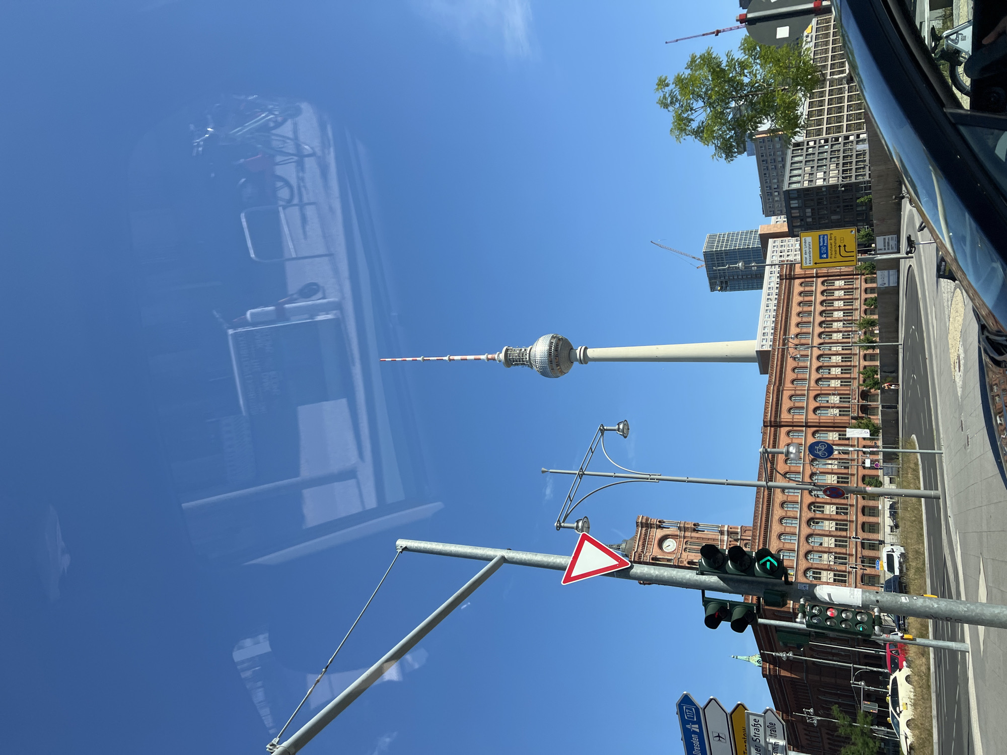 The image is a street-level view in Berlin, Germany, captured on a clear, sunny day. In the foreground, the hood and windshield of a dark car are visible. The main subjects of the image are the Fernsehturm (TV Tower) and the Rotes Rathaus (Red City Hall).
The Fernsehturm, a tall, slender tower with a silver sphere at its top and a red and white striped antenna, dominates the skyline. It is positioned towards the right side of the frame, seen against a bright blue sky.
The Rotes Rathaus, a large red brick building with a clock tower, is located on the left side of the image. It features numerous arched windows and a distinctive red facade. A triangular yield sign with a red border is prominently displayed on a metal pole in front of the Rotes Rathaus.
In front of the Rotes Rathaus, a traffic light is visible, showing a green arrow pointing to the right. Several other traffic signs are also present, including directional signs with German text. A street lamp with multiple lights stands near the traffic light.
In the middle ground, there are other buildings, including a modern high-rise under construction with cranes, and several other office or apartment buildings with various architectural styles. Trees, including a large green tree on the right side, are also present.
The street in front of the buildings appears to be a road with some traffic. A yellow taxi and other vehicles are visible. The image is partially obscured by reflections on the car's windshield, showing faint outlines of buildings and perhaps a structure with bicycles. The overall environment suggests an urban setting with a mix of historical and modern architecture.
