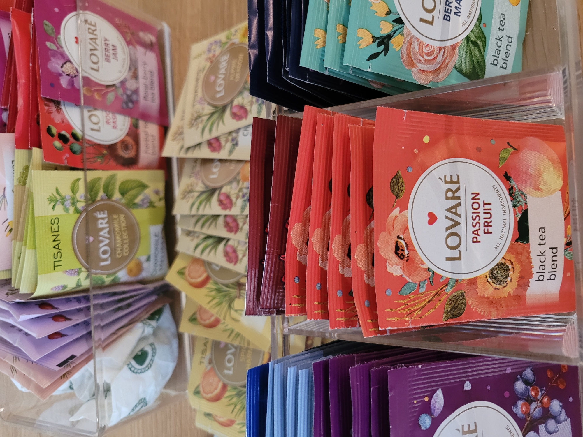 This media file shows a close-up, overhead view of a retail display of assorted tea sachets. The sachets are organized in clear plastic dividers, with multiple flavors visible. The display appears to be in a store setting, with the focus on the colorful packaging of the teas.
The subjects are the tea sachets themselves. They are predominantly in shades of pink, red, orange, yellow, green, and blue, each featuring floral or fruit imagery and branding. Visible text includes brand names like "LOVARÉ" and flavor descriptions such as "BERRY JAM," "ROCK RASP," "CHAMOMILE COLLECTION," "ALPINE MEADOW," "PASSION FRUIT," and "black tea blend." Some sachets also have text indicating "ALL NATURAL INGREDIENTS."
The scene is a retail display, likely in a grocery store or specialty shop. The lighting suggests an indoor environment, possibly during daylight hours. There are no people or specific weather conditions depicted. The setting is organized and clean, highlighting the product for sale. The overall impression is one of variety and appeal, showcasing the different flavors and designs of the tea offerings. The location context of Kyiv, Ukraine, is not directly evident from the visual information but is provided as additional metadata.