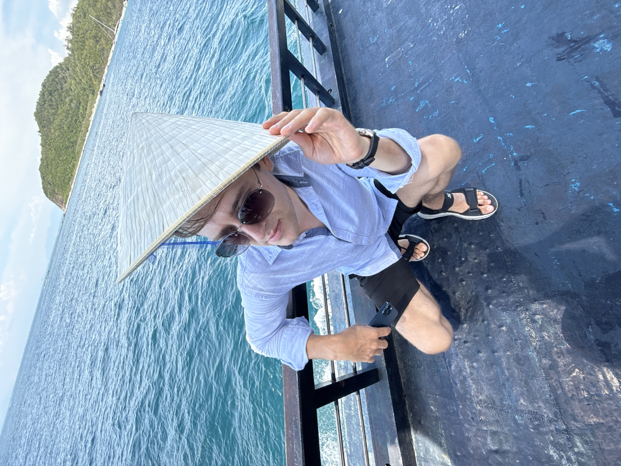 A man kneels on a dark-colored surface with ocean water and a green island visible in the background. The man is wearing a light blue button-down shirt, dark shorts, and sandals. He also wears sunglasses and a traditional Asian conical hat. He holds a phone in his left hand and adjusts his hat with his right. The dark-colored surface appears to be a deck with dark metal railings. The ocean is blue with visible waves, and the island is lush green with trees. The location is Nha Trang Ward, Vietnam.