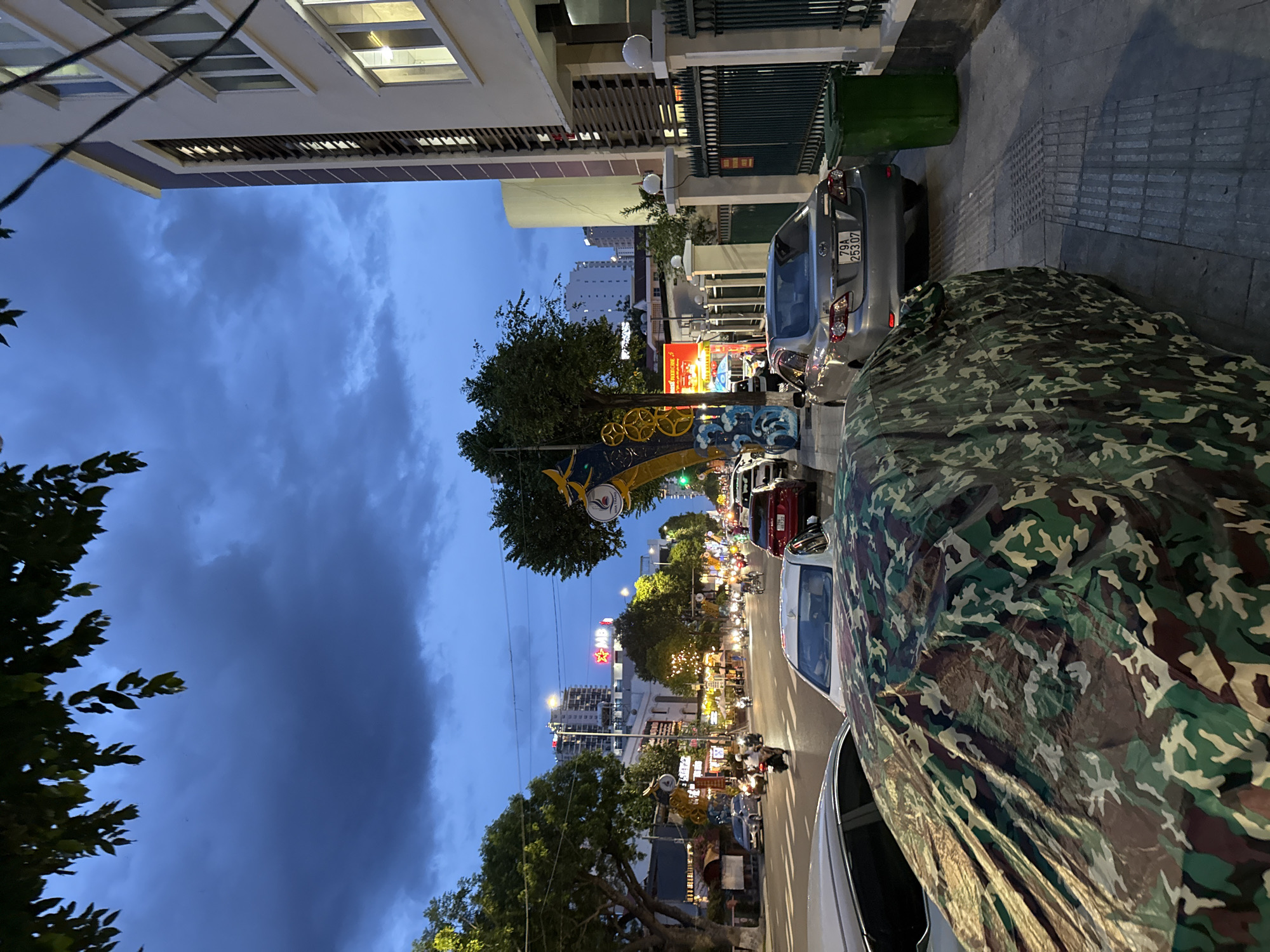 The image depicts a street scene in Nha Trang, Vietnam, during dusk or early evening. The sky is a deep blue with dramatic clouds, suggesting an approaching storm or recent rain. The street is lined with buildings, trees, and parked cars, with active traffic including motorcycles and cars. A prominent building on the right side of the image is a light-colored structure with multiple windows and dark vertical accents. A black metal fence runs along the sidewalk in front of this building. A green trash bin is visible next to the fence. In the foreground, two cars are parked on the left side of the street, one of which is covered with a camouflage car cover. The street itself is paved, with clear lane markings. Businesses with illuminated signs are visible along the street, indicating a commercial area. Trees with lush green foliage are interspersed along the street, casting shadows. The overall impression is of a bustling urban environment at the transition from day to night.