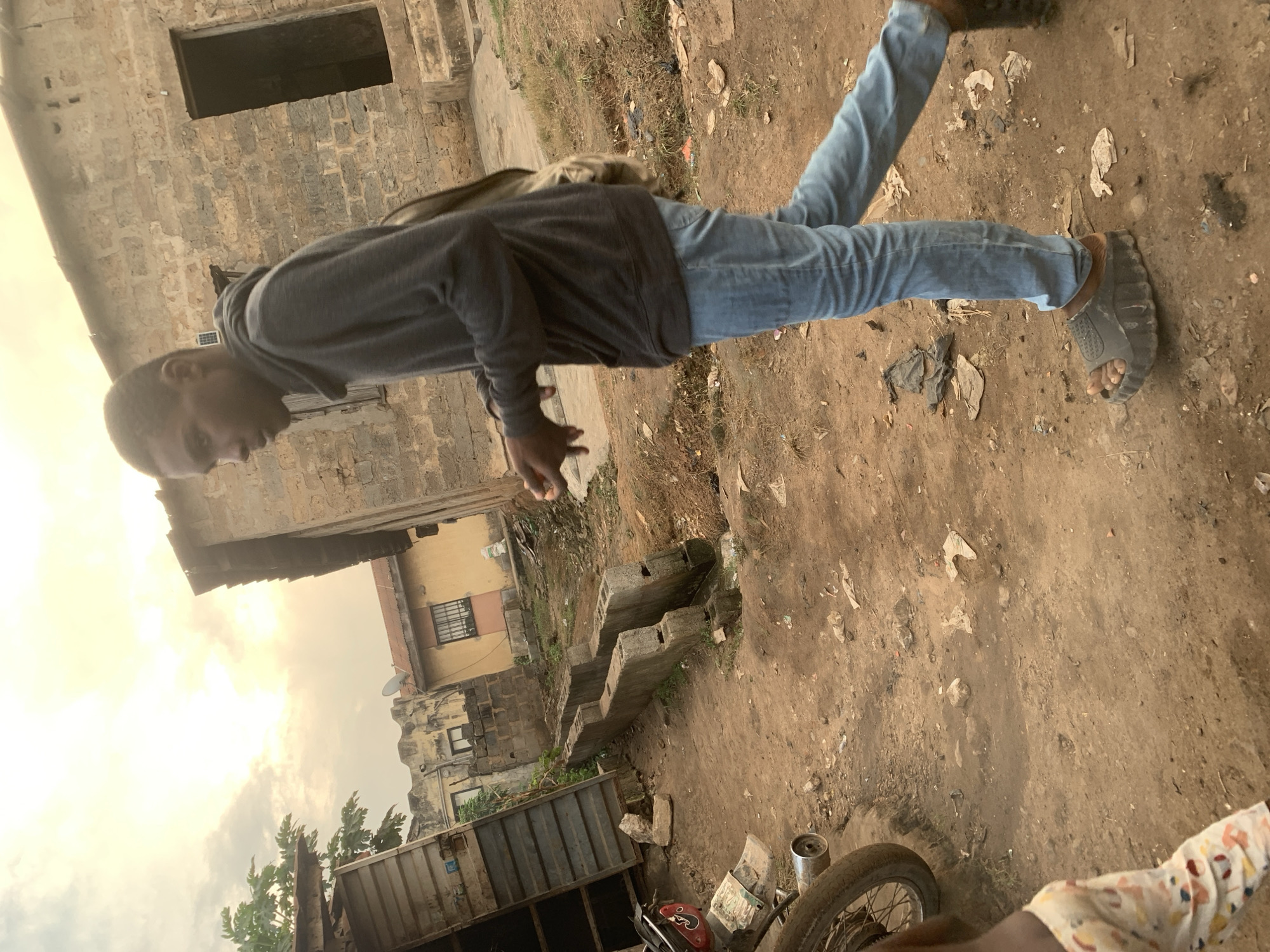 A young Black male walks across a dirt path in Shagamu, Nigeria. The man is wearing a dark gray hooded sweatshirt, light blue jeans, and dark gray sandals. He has a bag slung over his right shoulder. He is walking towards the right side of the frame, with his head turned slightly to the left, looking at something off-camera. The environment appears to be an outdoor area with several buildings in the background, some of which are made of cinder blocks and others with yellow painted exteriors. There is a motorcycle parked to the left of the frame. The ground is covered in dirt, with some debris and vegetation visible. The sky is overcast with soft, diffused light, suggesting it may be early morning or late afternoon.