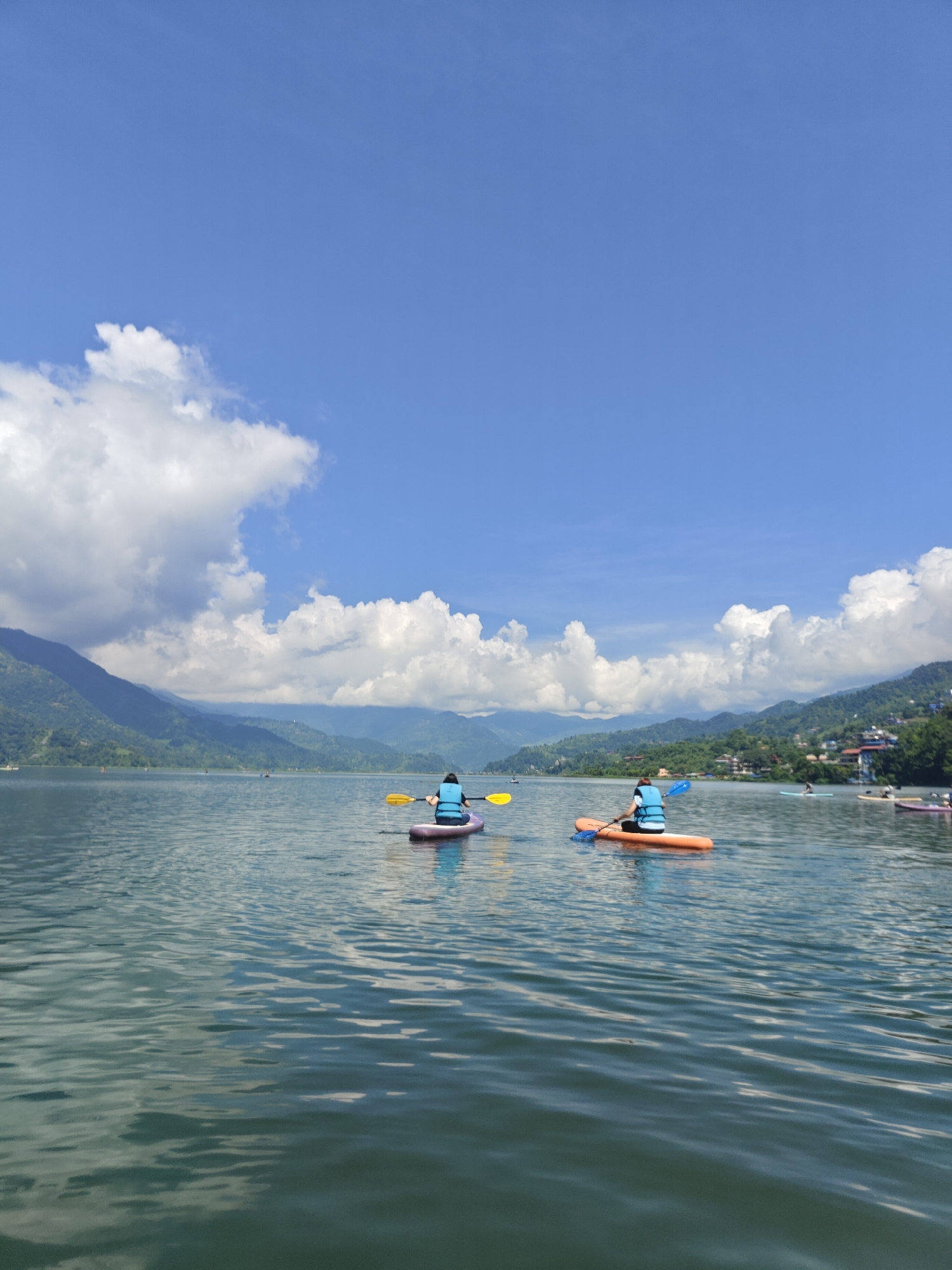 This image depicts a serene scene on Phewa Lake in Pokhara, Nepal. Two individuals are actively engaged in watersports, with one person paddling a purple kayak and another on an orange paddleboard. Both are wearing blue life vests, indicating safety precautions. The lake's surface is calm, reflecting the clear blue sky and scattered white clouds, with majestic, tree-covered hills forming a picturesque backdrop. In the distance, the city of Pokhara is visible, nestled along the shore, with hints of buildings and further mountainous terrain. The overall atmosphere is one of tranquility and outdoor recreation amidst the stunning natural beauty of Nepal.