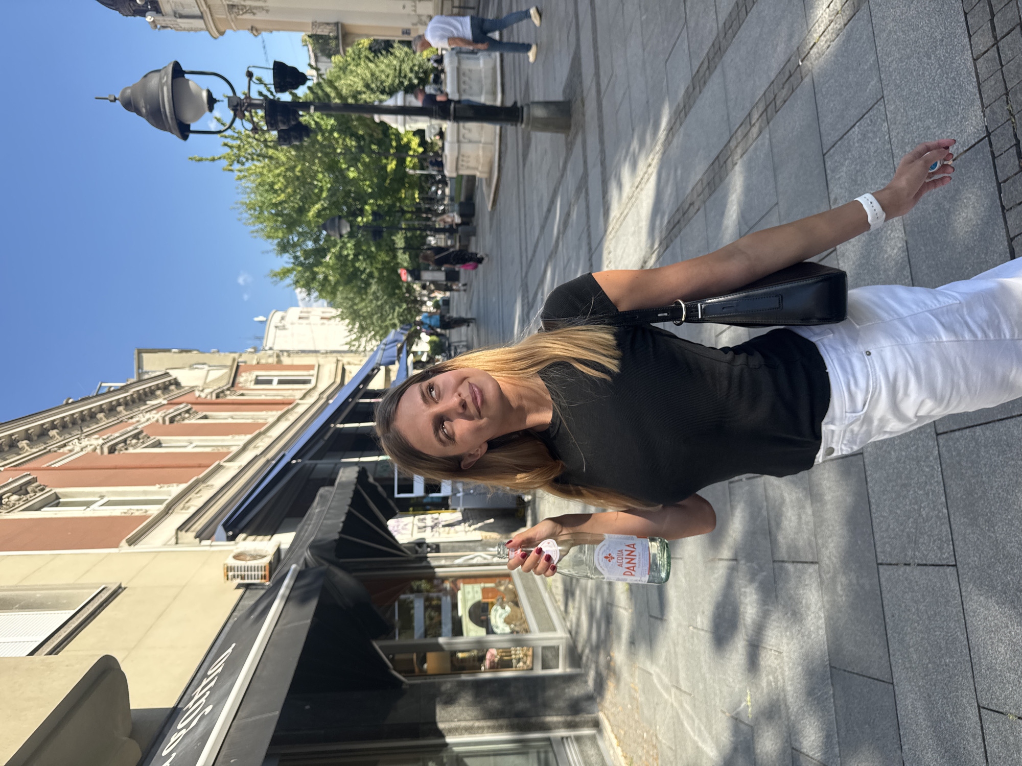 A young woman with blonde hair is holding a bottle of Acqua Panna water as she walks down a paved pedestrian street. She is wearing a black t-shirt and white pants, and carrying a black handbag. She is looking up and to her right, with a slight smile. The street is lined with buildings, some with awnings over shop fronts. There are trees and lampposts along the street, and other people can be seen walking in the background. The scene appears to be in Belgrade, Serbia, possibly Knez Mihailova Street, which is a well-known pedestrian zone.