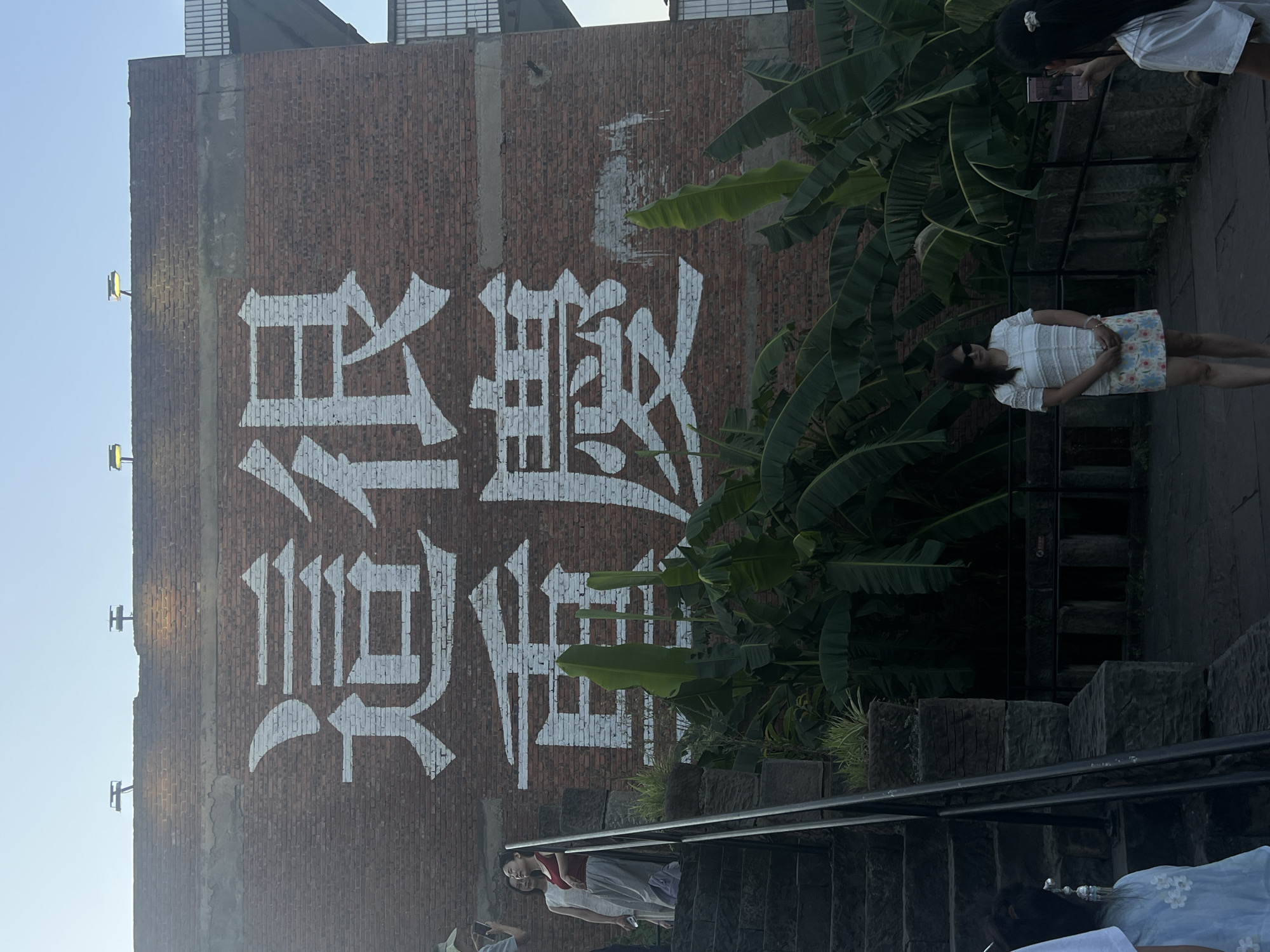 The image captures a wall in Nan'an District, China, adorned with large white Chinese characters painted in a bold, calligraphic style. The characters translate to "This is Chongqing," a phrase often used to express pride in the city. The brick wall serves as a canvas, with the white paint starkly contrasting against the reddish-brown bricks. Above the wall, the sky is a clear, light blue, suggesting a daytime scene. Two modern-looking light fixtures are mounted on the top edge of the wall.
In the foreground, a set of stone stairs ascends to the left, with metal railings. Several people are present in the scene. On the stairs, a woman in a red top and gray skirt stands, looking upwards and slightly to the left. Another woman in a white dress stands beside her. Further down the stairs, a person wearing a light blue garment is partially visible. To the right of the stairs, a woman wearing sunglasses, a white lacy top, and a floral skirt is standing, facing the viewer. To her right, another person is holding up a smartphone, appearing to take a photograph. Lush green foliage, including large banana leaves, grows around the base of the wall and along the pathway. In the background, a modern structure with visible grids is present to the right of the wall. The overall setting appears to be an outdoor public space, possibly a park or a historical site within the city.