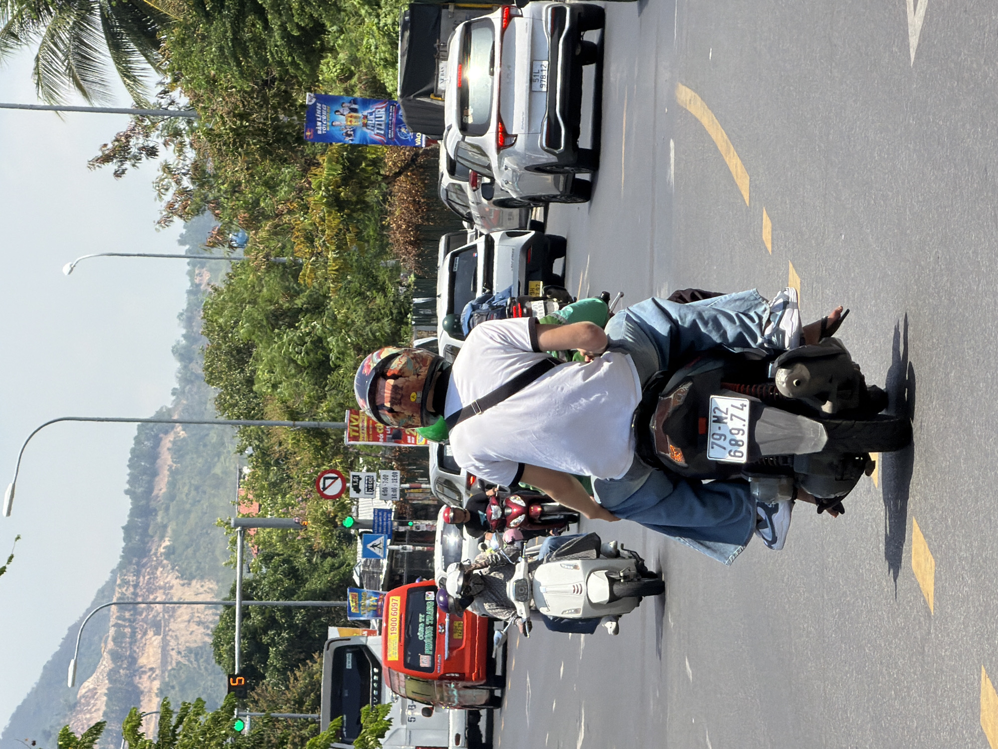 The image depicts a street scene in Nam Nha Trang Ward, Vietnam, on a sunny day. A convoy of motorbikes and cars are navigating a multi-lane road. In the foreground, a person on a motorbike, wearing a white t-shirt and blue jeans, is facing away from the camera. They have a black helmet with a colorful pattern and are riding a black motorbike with a white license plate that reads "79-M2 689.74". Behind this motorbike, another person is also on a motorbike, partially visible.
Further down the road, a white scooter is carrying two individuals, both wearing helmets. To the left, a red van with the number "1900 6097" on its side is stopped, with text in Vietnamese visible on its doors. Several other vehicles, including white cars and motorbikes, are also present on the road.
Traffic lights, including a green light, are visible above the road, along with street signs and poles. Trees and vegetation line the sides of the road, and in the background, a hilly landscape with exposed earth and trees is visible. The road surface is asphalt with yellow and white markings. The overall impression is a busy urban street with typical traffic conditions.